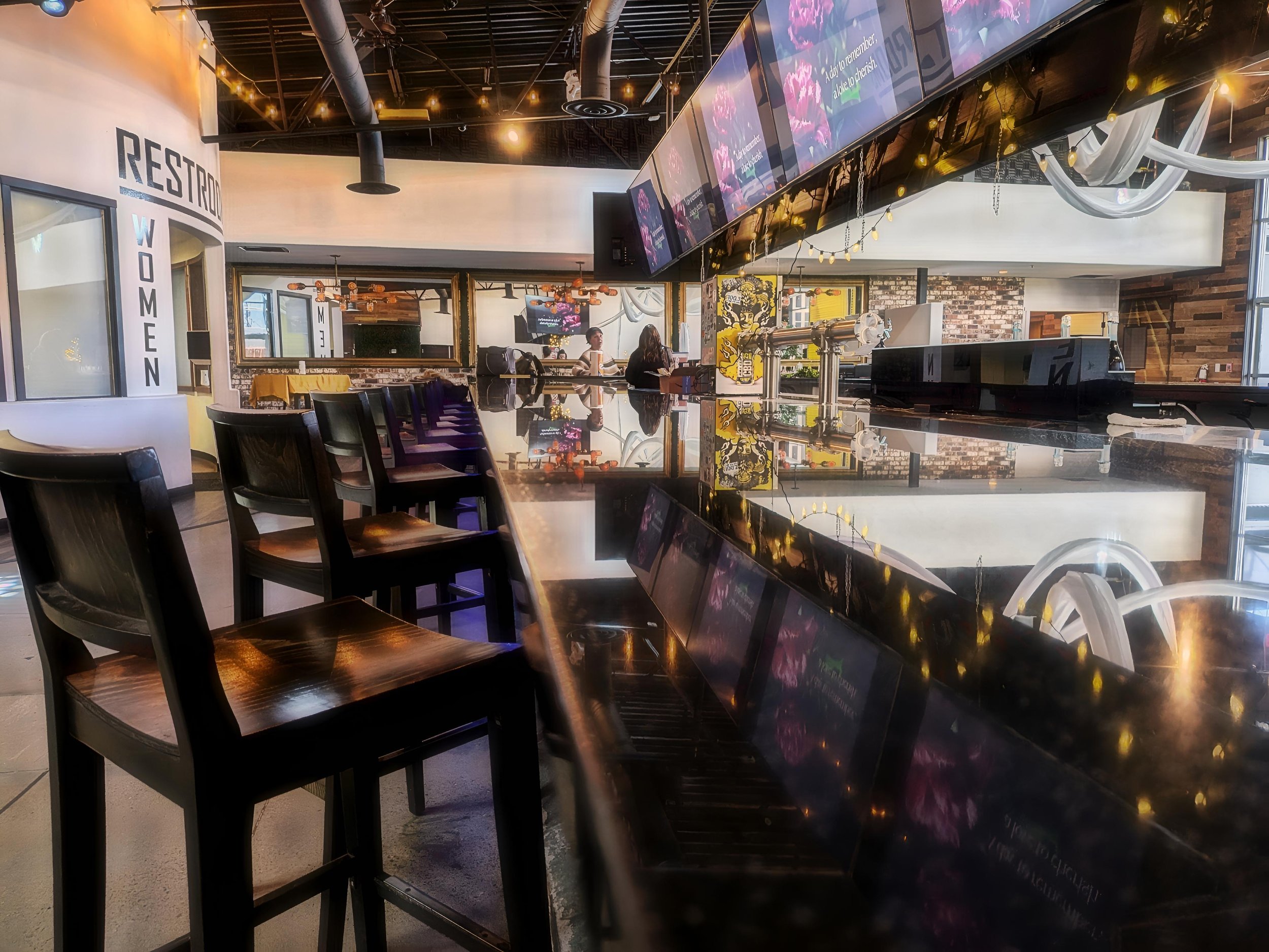 Inside a modern cafe with a bar-top counter, wooden chairs, and a large digital menu screen hanging above. There are people at the counter and church-style windows allowing natural light. The decor includes brick walls and hanging string lights.