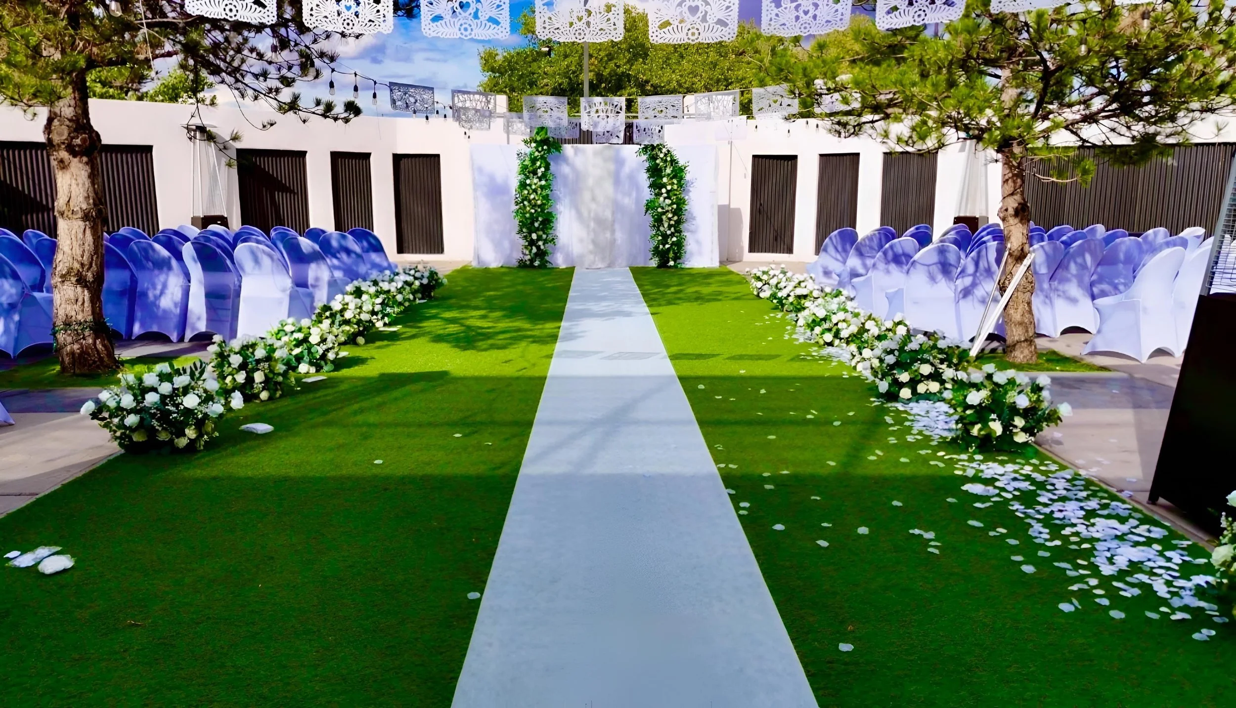 Outdoor wedding ceremony setup with a white aisle runner, floral decorations, white chairs with covers, and green trees under a sunny sky.
