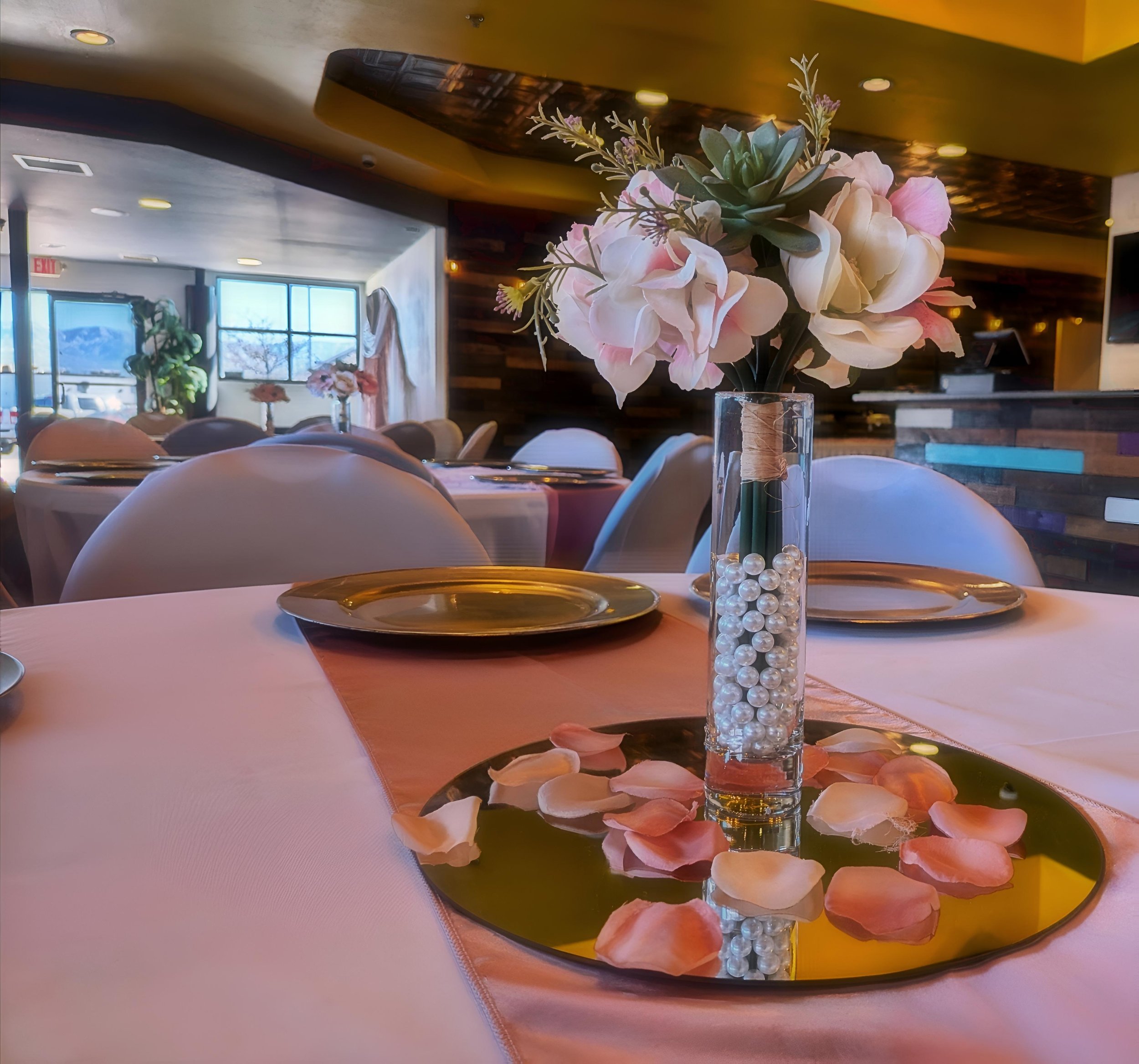 A table decorated with a flower arrangement in a glass vase, surrounded by rose petal confetti on a round mirrored tray, in a softly lit restaurant or banquet hall.