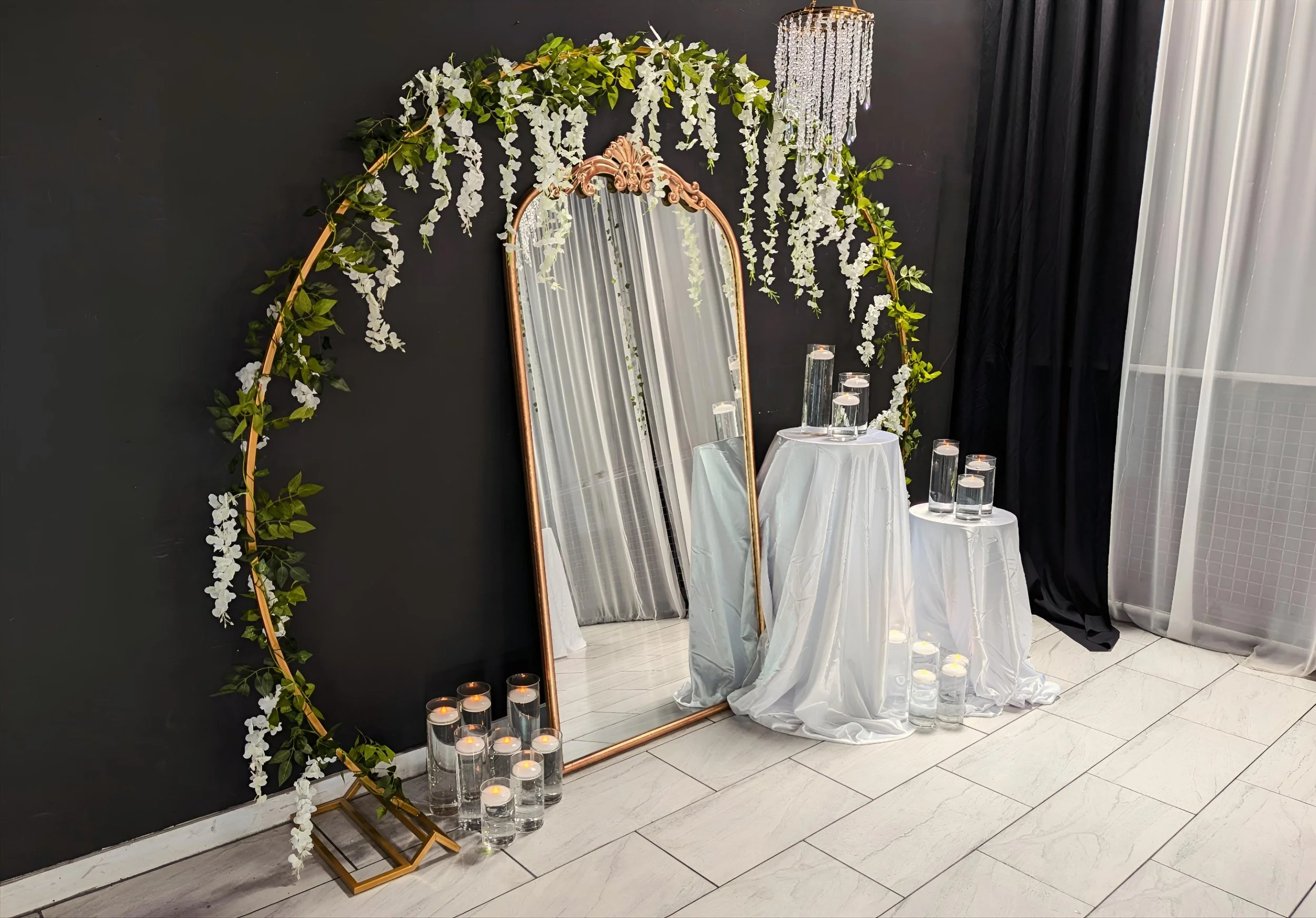 Decorative setup with a tall mirror, floral arch, candles, and small tables with vases in an elegant interior