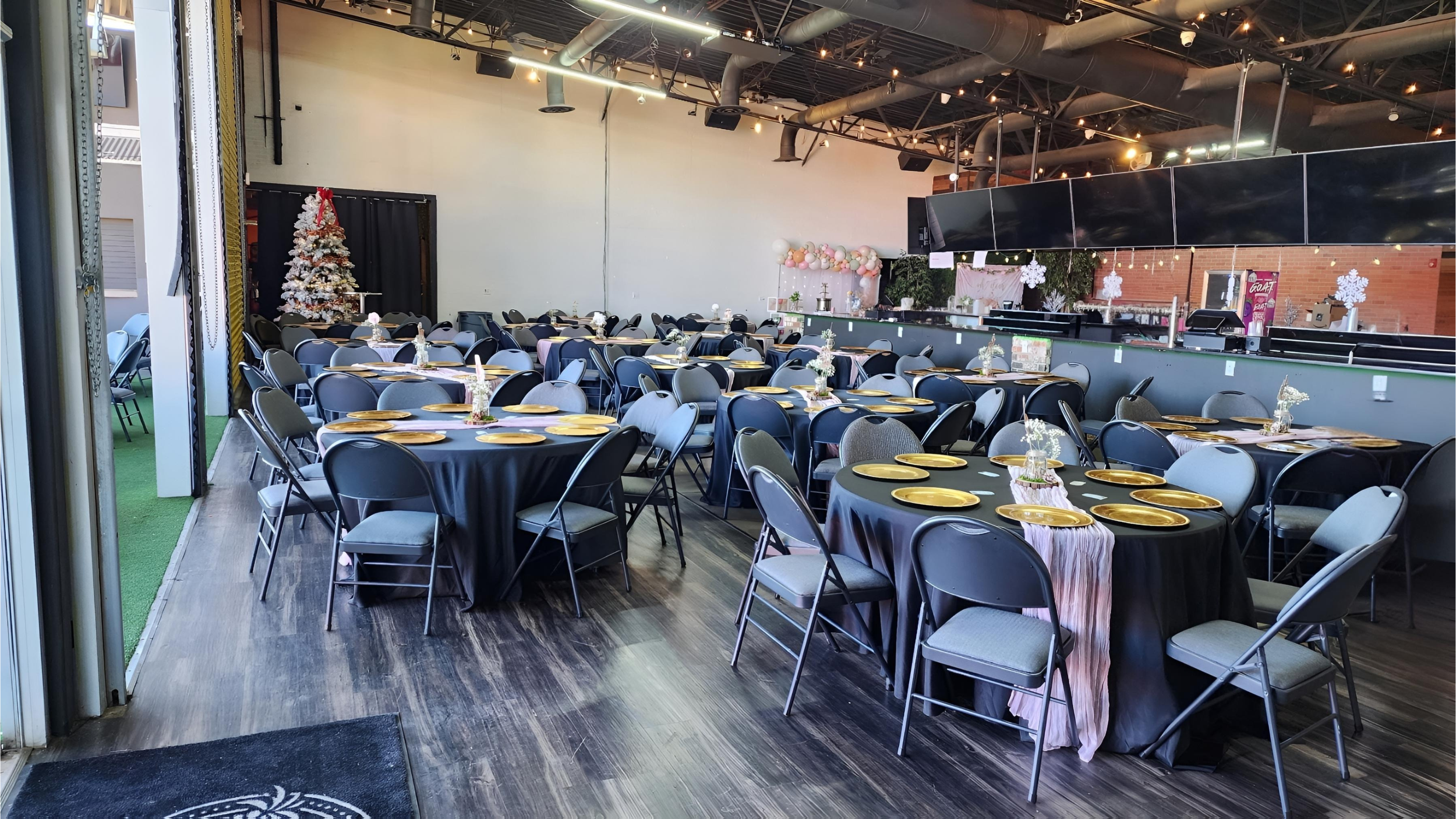 Indoor event space set up with round tables covered in black tablecloths, decorated with small floral centerpieces, and gold plates arranged for a celebration, featuring a Christmas tree with white ornaments and ribbons, and pink and white balloon de
