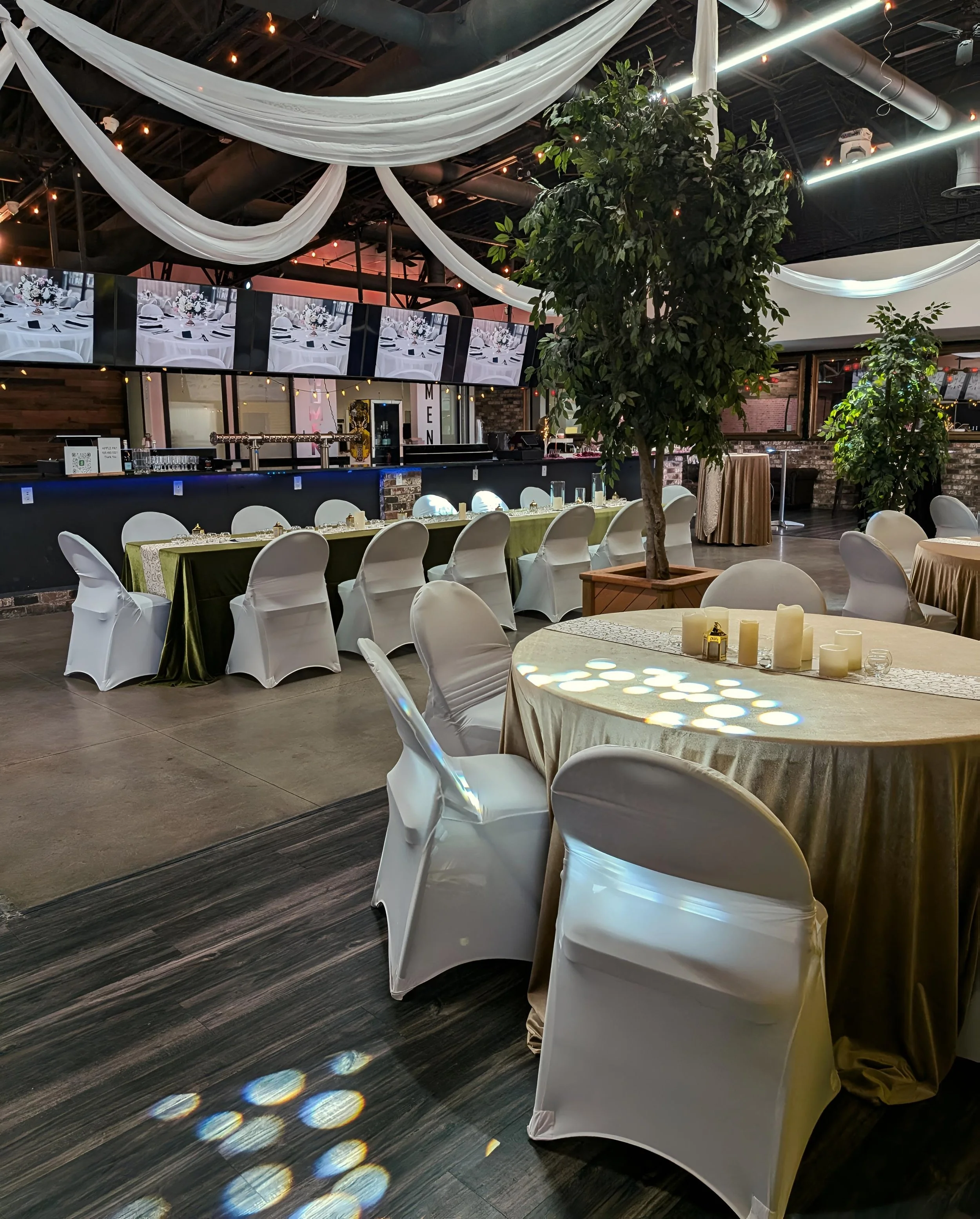 Elegant event venue decorated with white chair covers, draped fabric, and green plants, featuring long tables with candles and a bar area with multiple monitors displaying a table setting.