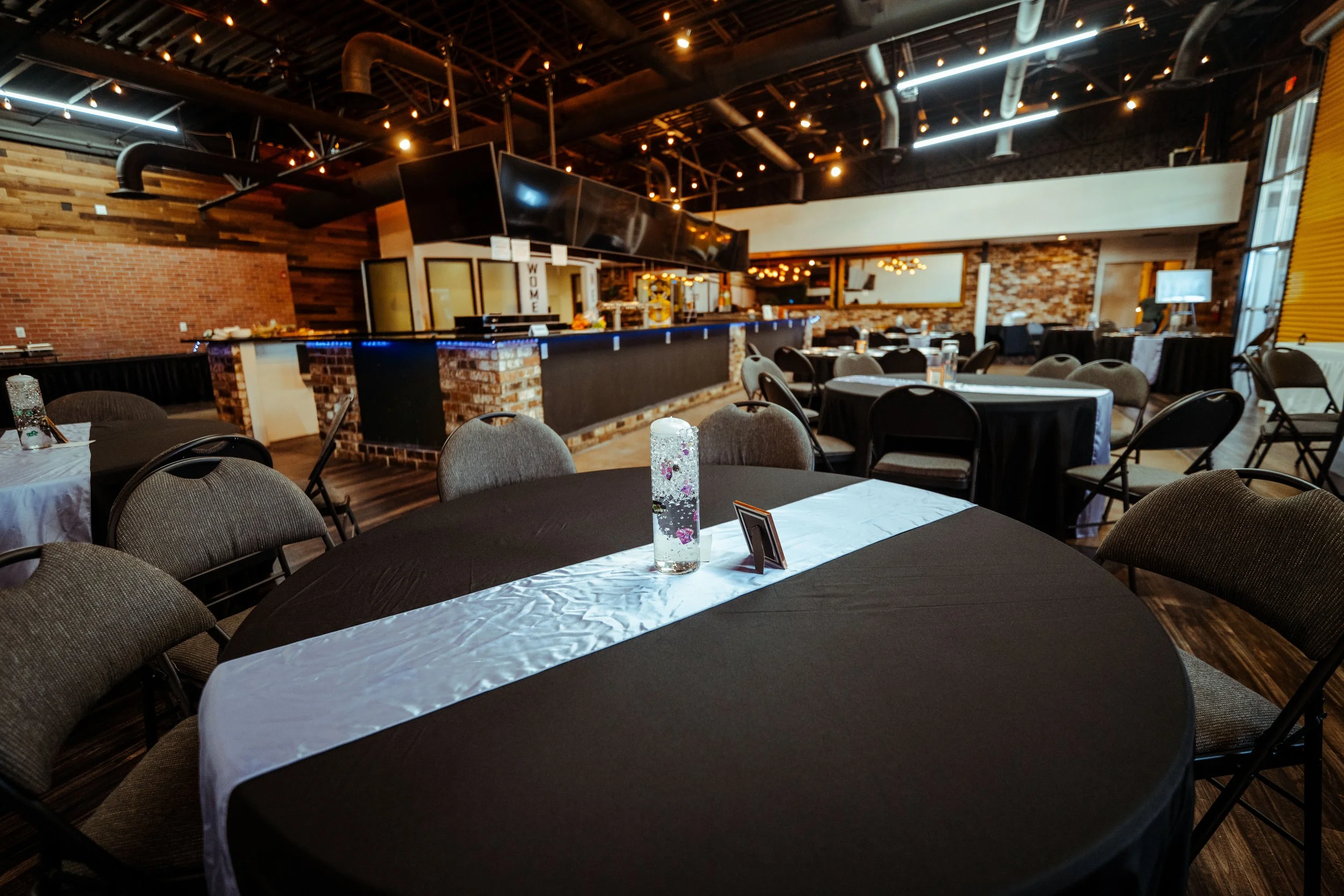 Interior of a modern event space with round tables covered in black tablecloths and gray upholstered chairs, some with photo frames and a decorative candle holder on the table in the foreground, brick and wood accent walls, and a bar area with a bric