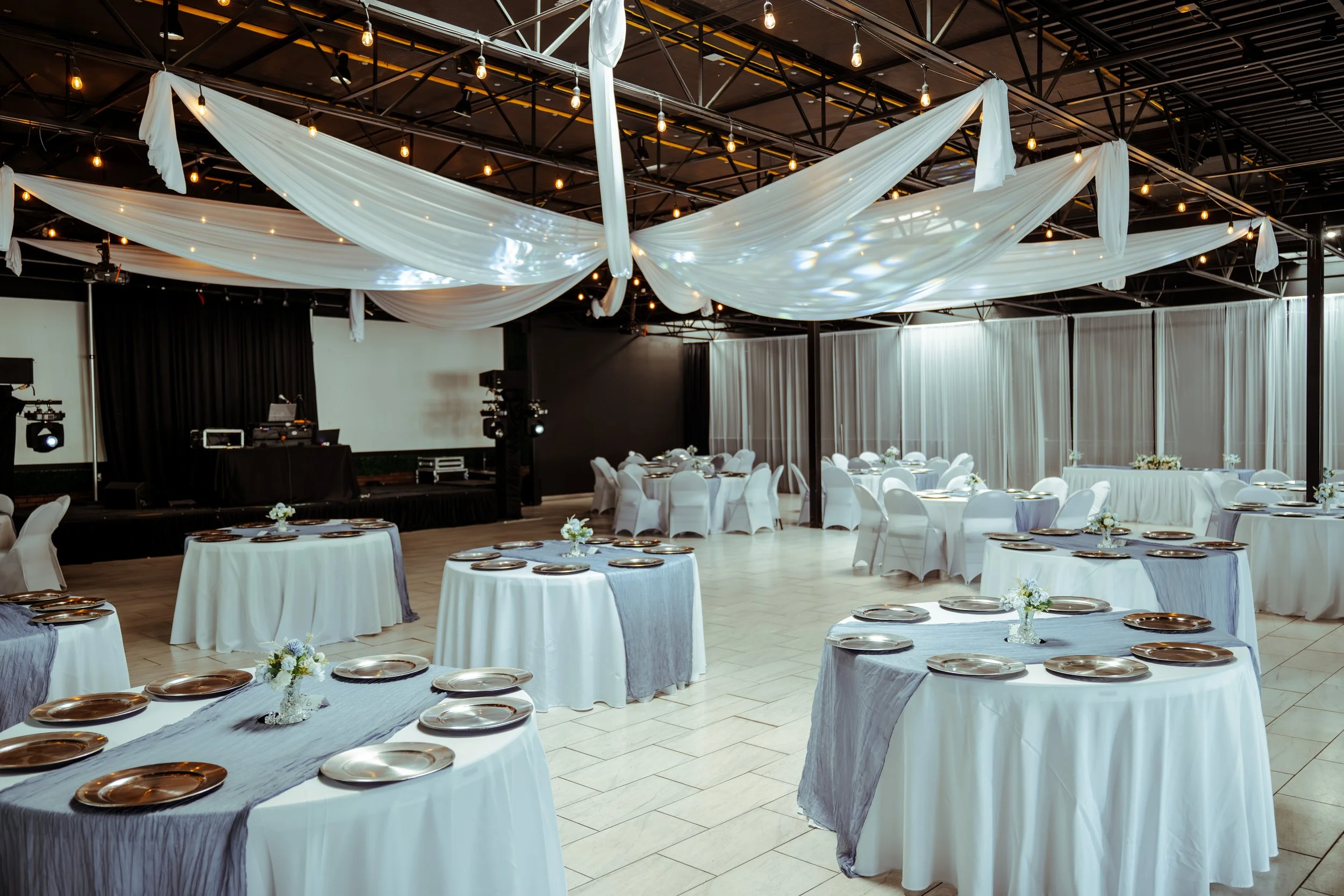 Elegant banquet hall decorated with white tablecloths, silver plates, and small floral centerpieces. White drapes and string lights adorn the ceiling. A stage with DJ equipment and lighting is visible at the back.
