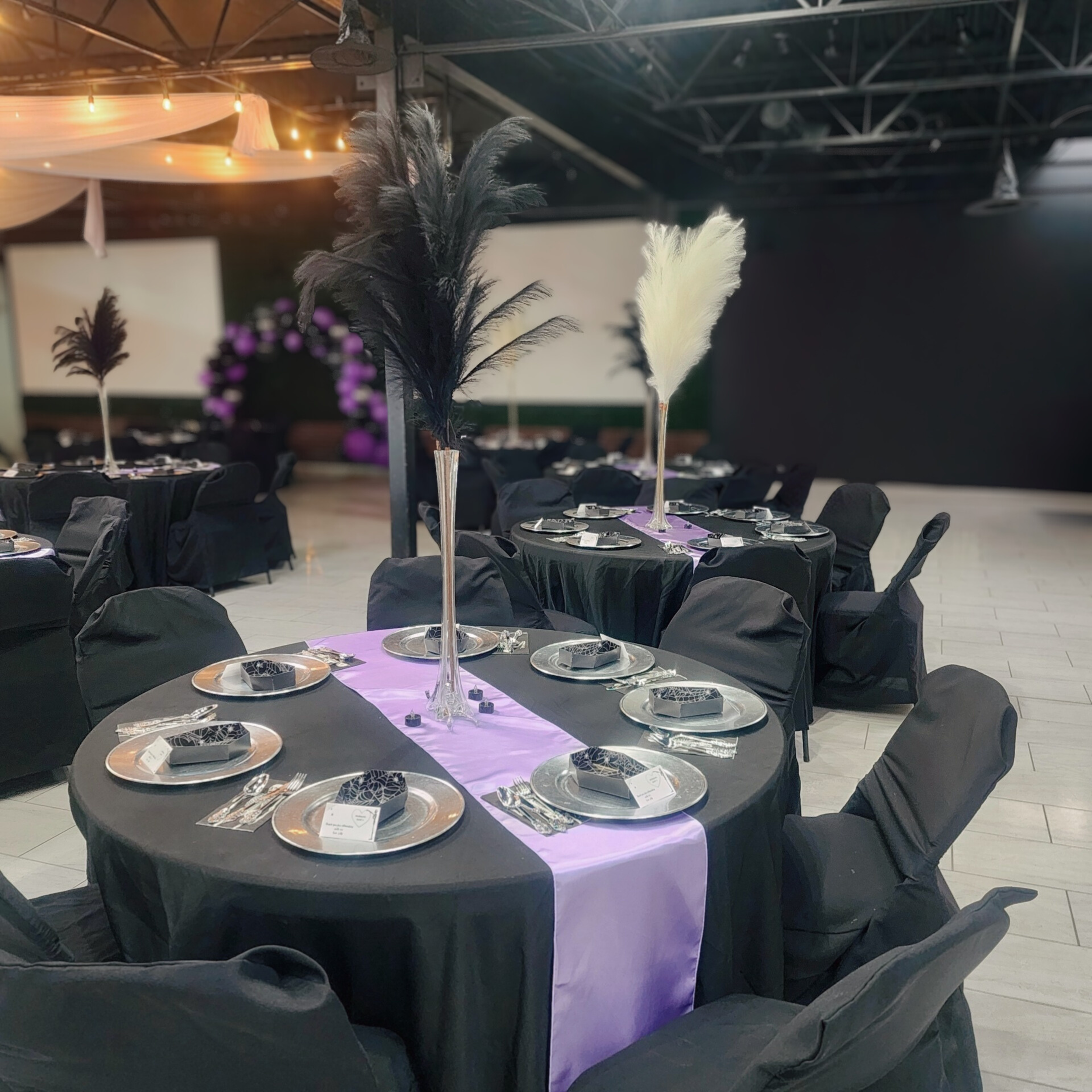 Elegant event table decorated with black tablecloths, purple table runners, tall black and white feather centerpieces, and black chair covers, set for a formal dinner.