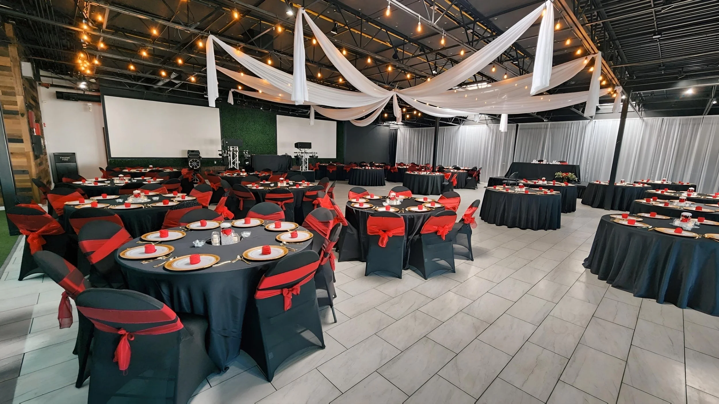 Event hall decorated for celebration with black tables covered with black tablecloths, red and black chairs tied with red ribbons, and place settings with plates, glasses, napkins, and candles. White drapery and string lights adorn the ceiling, and a