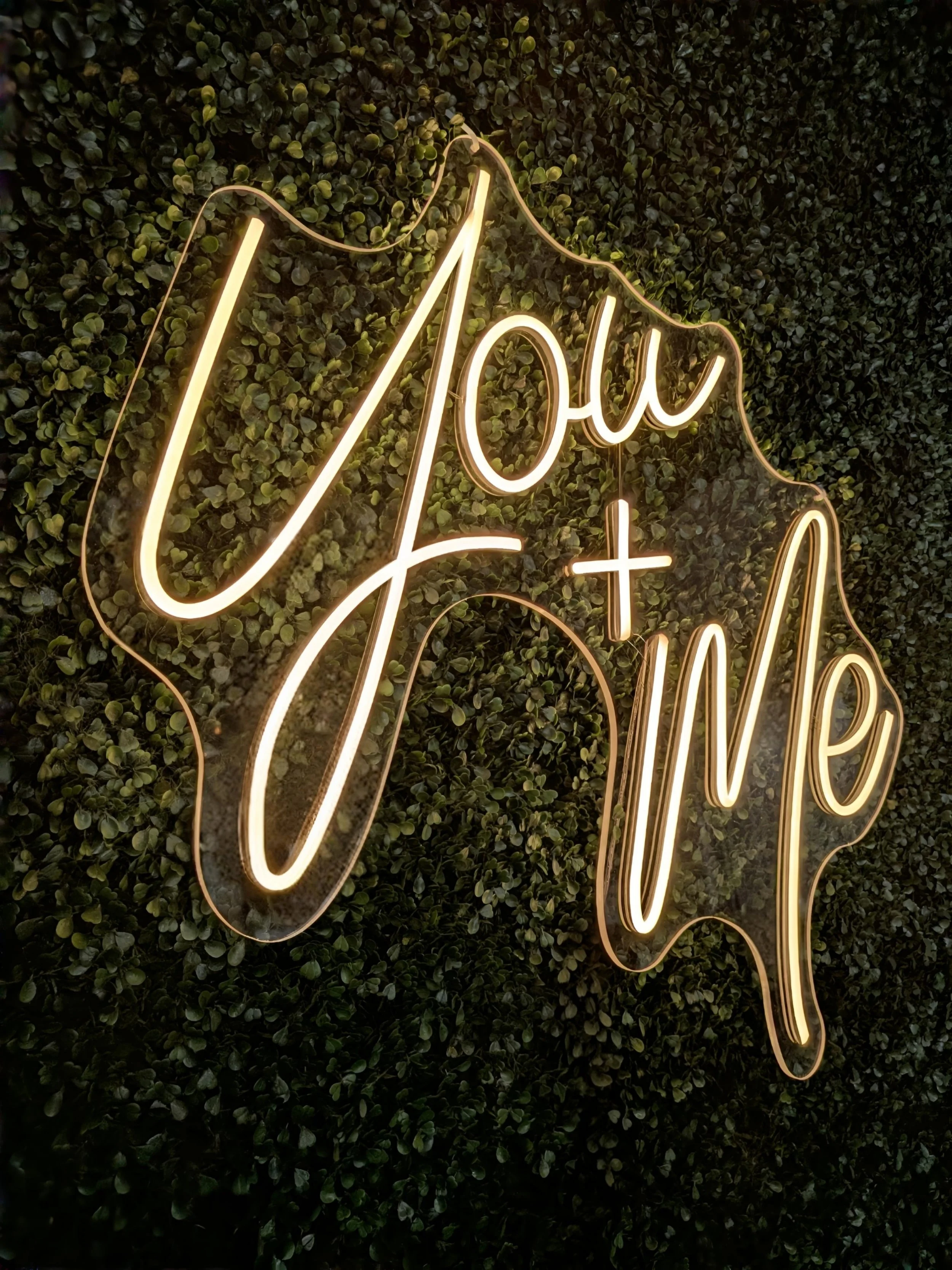 Neon sign saying 'U You + Me' on a leafy green wall.