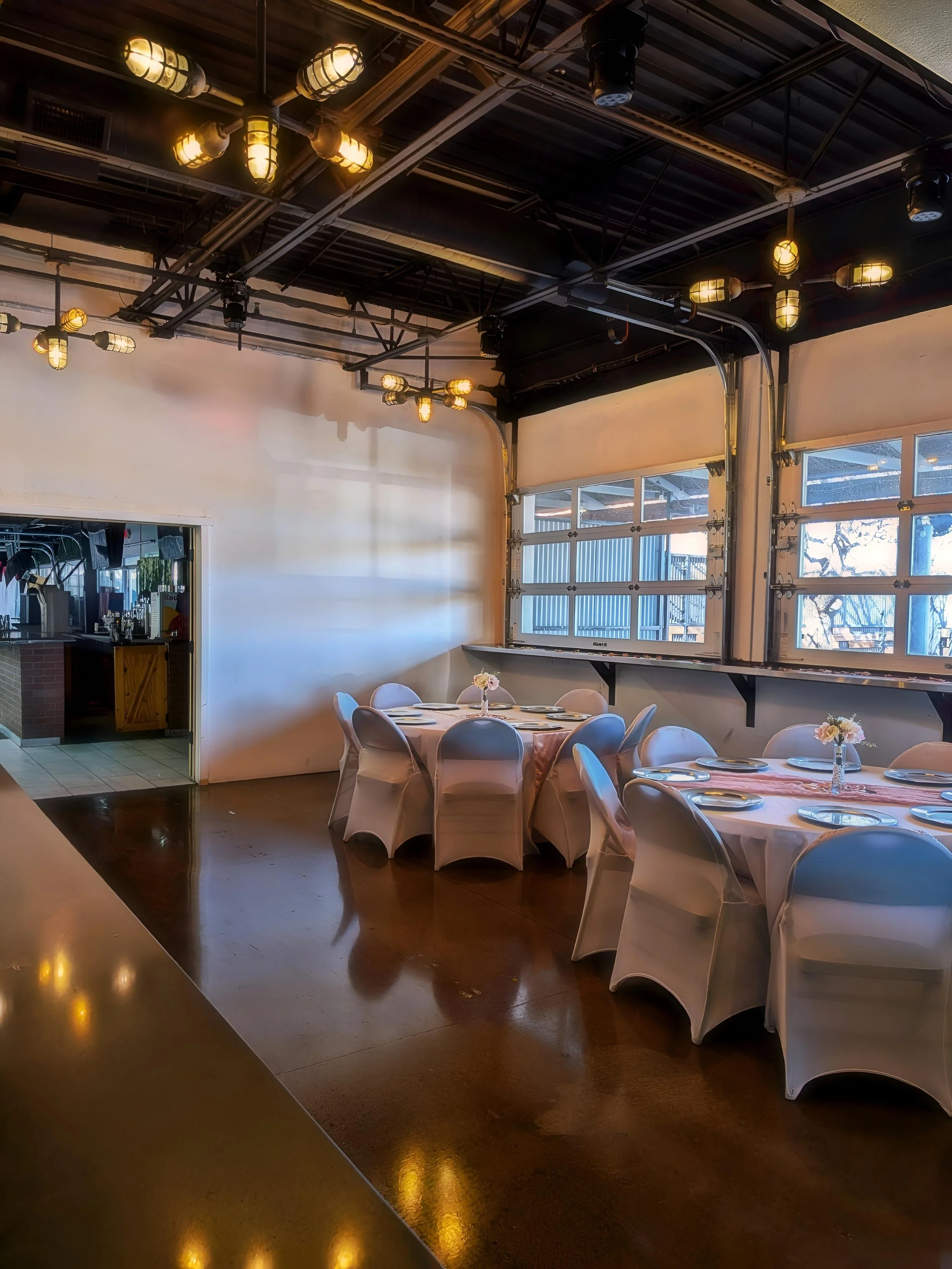 Empty restaurant with round tables covered in white tablecloths, decorated with small flower arrangements, and surrounded by white chairs. Large garage-style windows allow natural light, with exposed ceiling pipes and lighting fixtures.