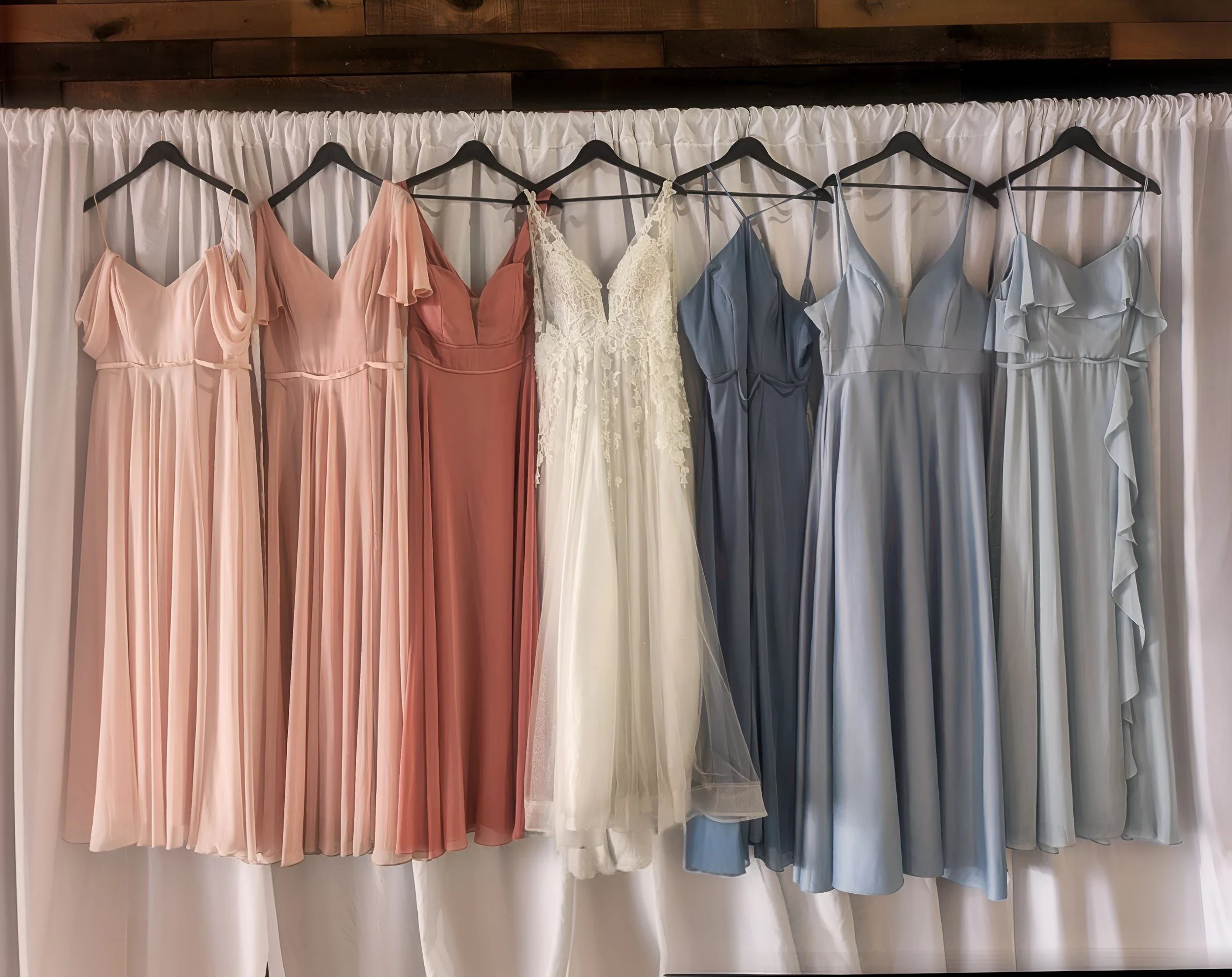 A selection of seven bridesmaid dresses hanging on black hangers against a white curtain backdrop. The dresses are in soft pastel shades, from blush pink to blue, with a white bridal gown in the middle.
