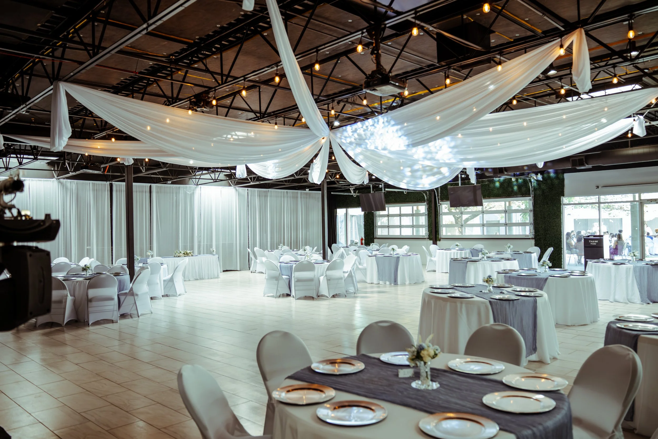 Elegant banquet hall decorated with white drapery and string lights, with round tables set with white plates and flowers, ready for a wedding or formal event.