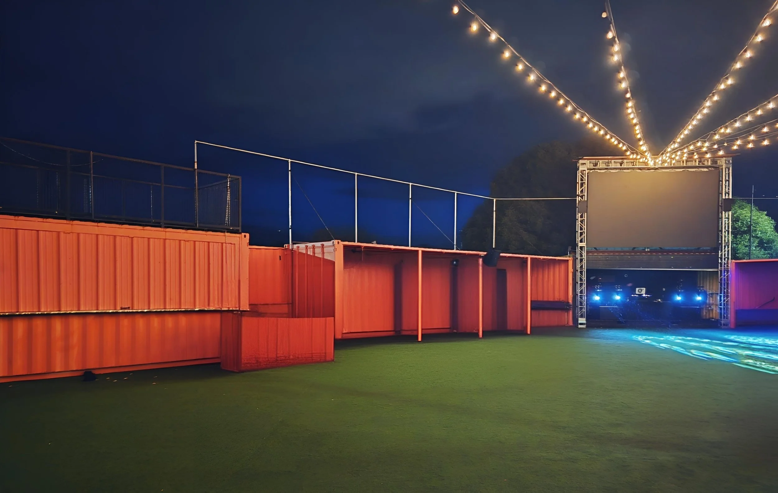 An outdoor event space at night with string lights overhead, a large blank screen, orange shipping containers, and blue lighting in the background.