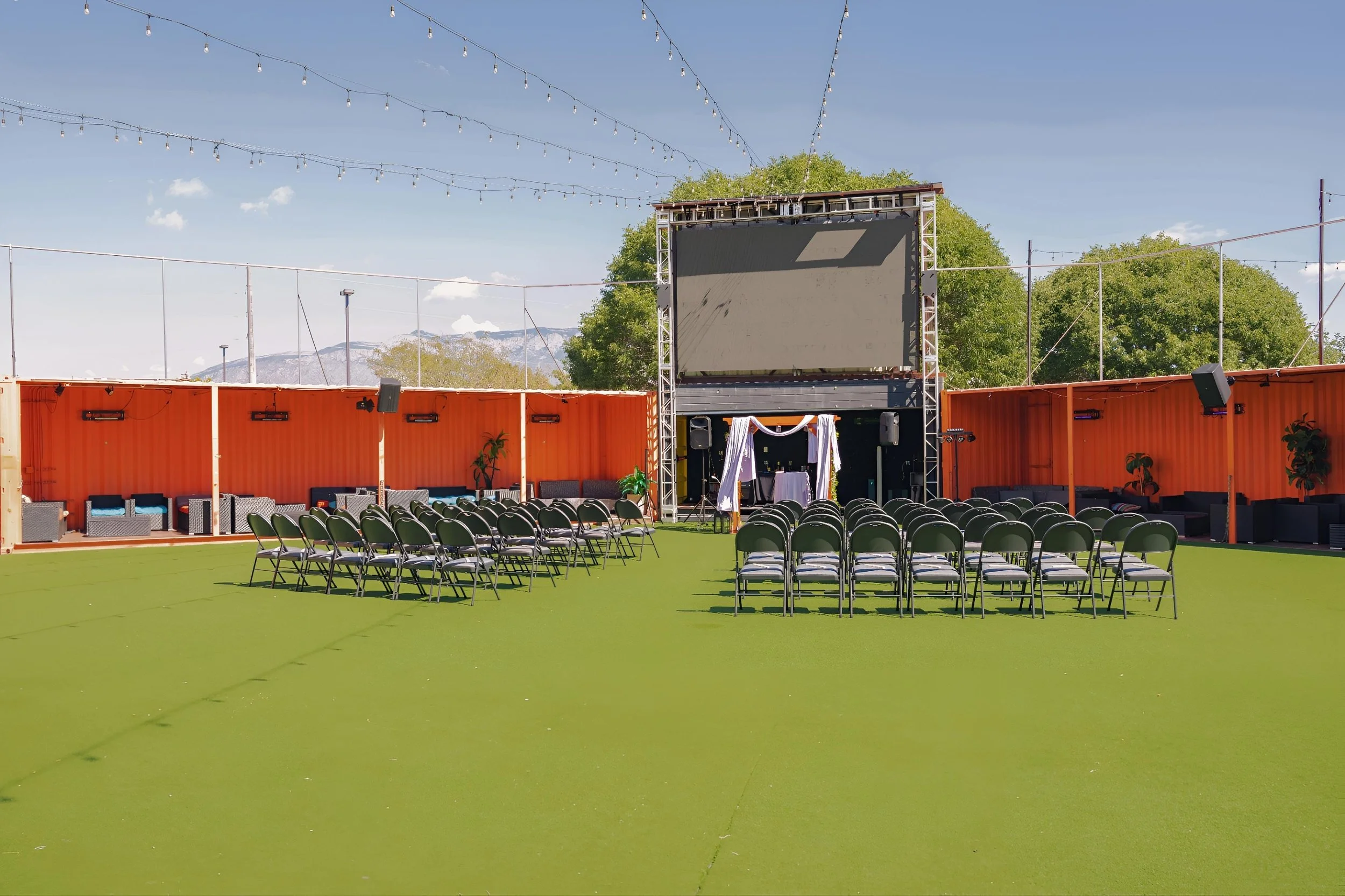 An outdoor event space with a large screen, rows of chairs, string lights overhead, and a small stage decorated with white drapes and greenery, surrounded by high orange fencing and seating areas.