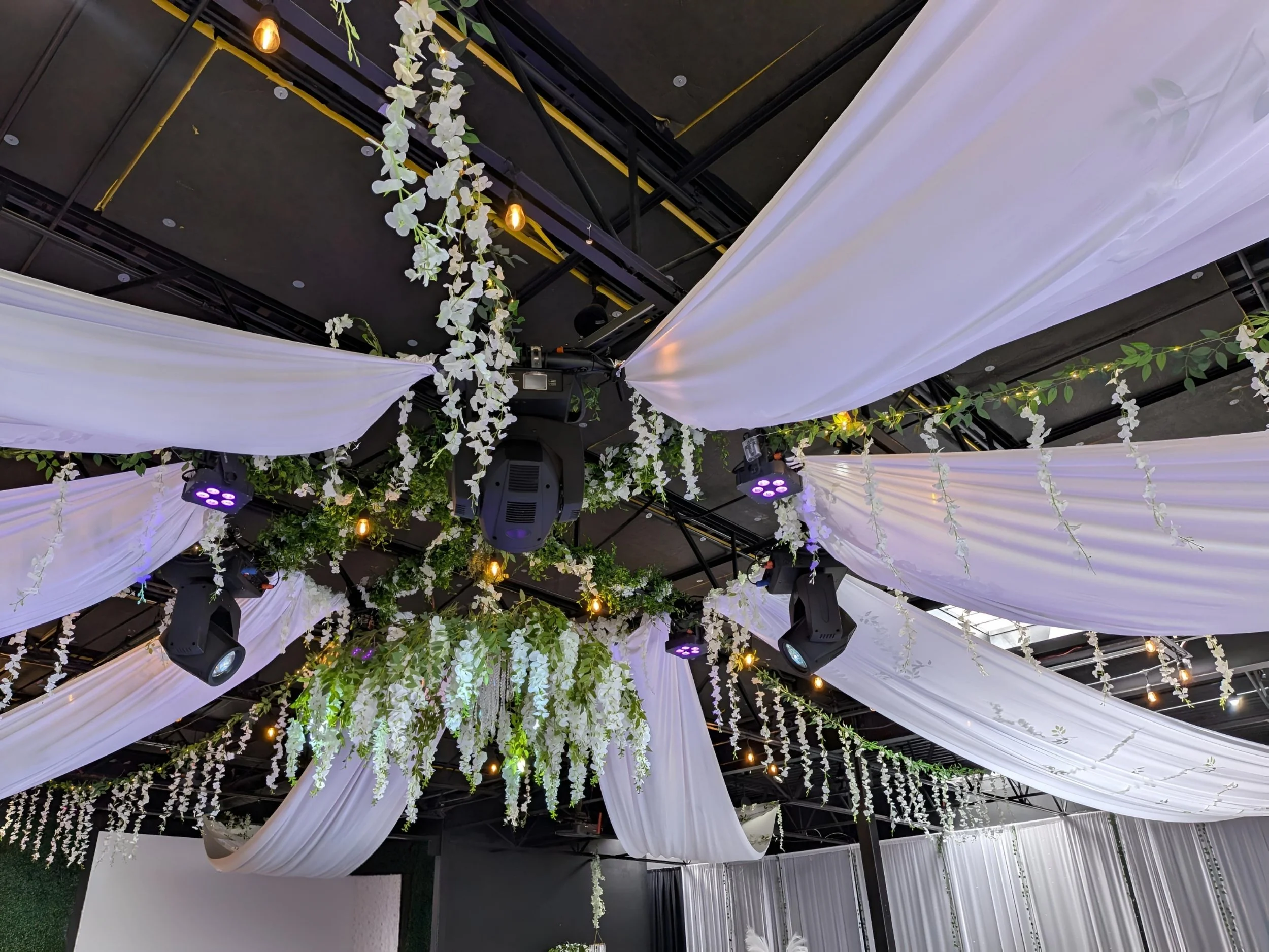 Ceiling decorated with white draped fabric, hanging white flowers, green foliage, and framed by black metal beams, with stage lighting.