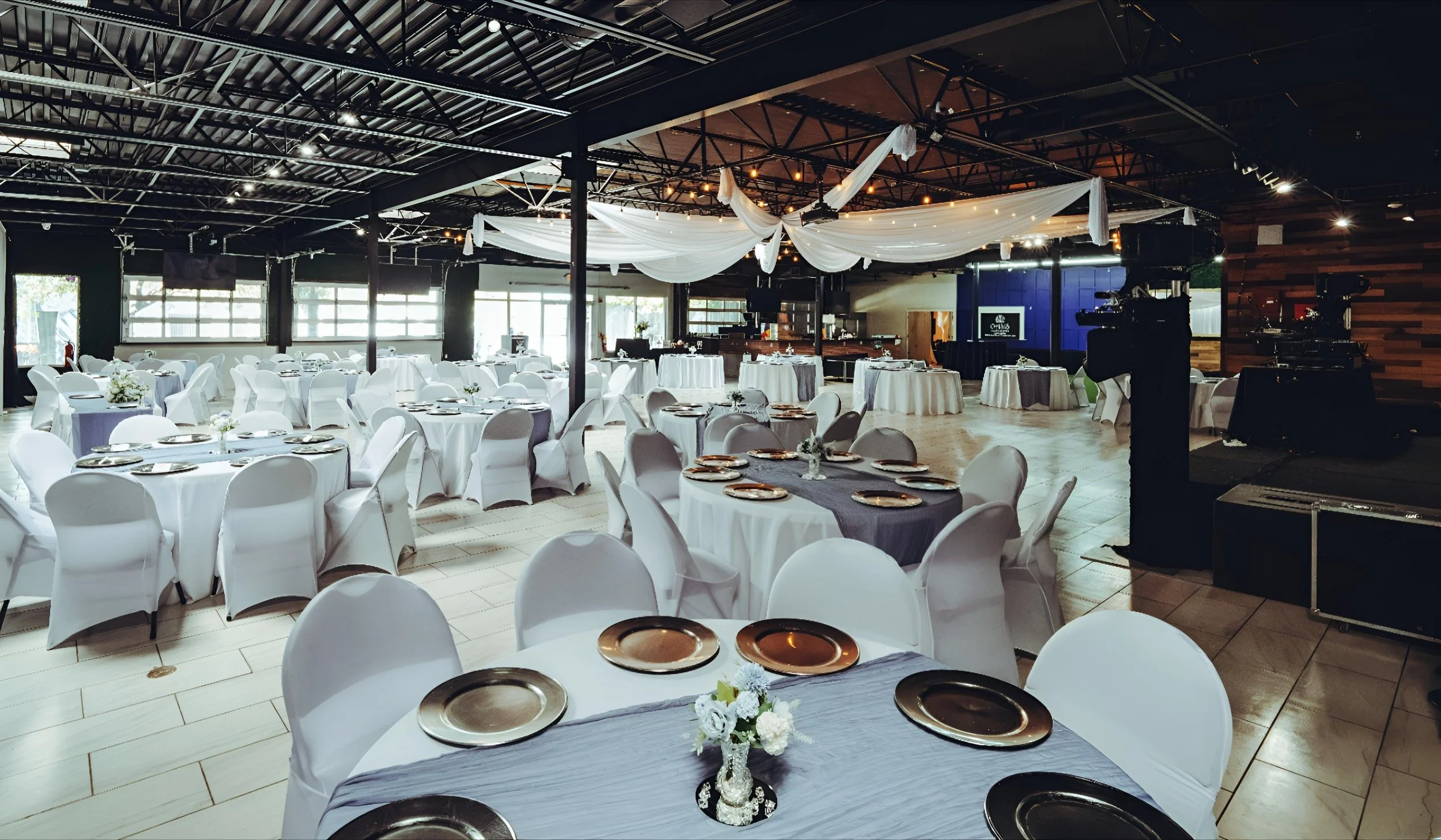 Wedding reception setup with round tables covered in white tablecloths, white chair covers, and gold-rimmed plates. Decor includes small floral centerpieces and draped white fabric hanging from the ceiling. The venue has large windows, wooden accents, and professional lighting and sound equipment.