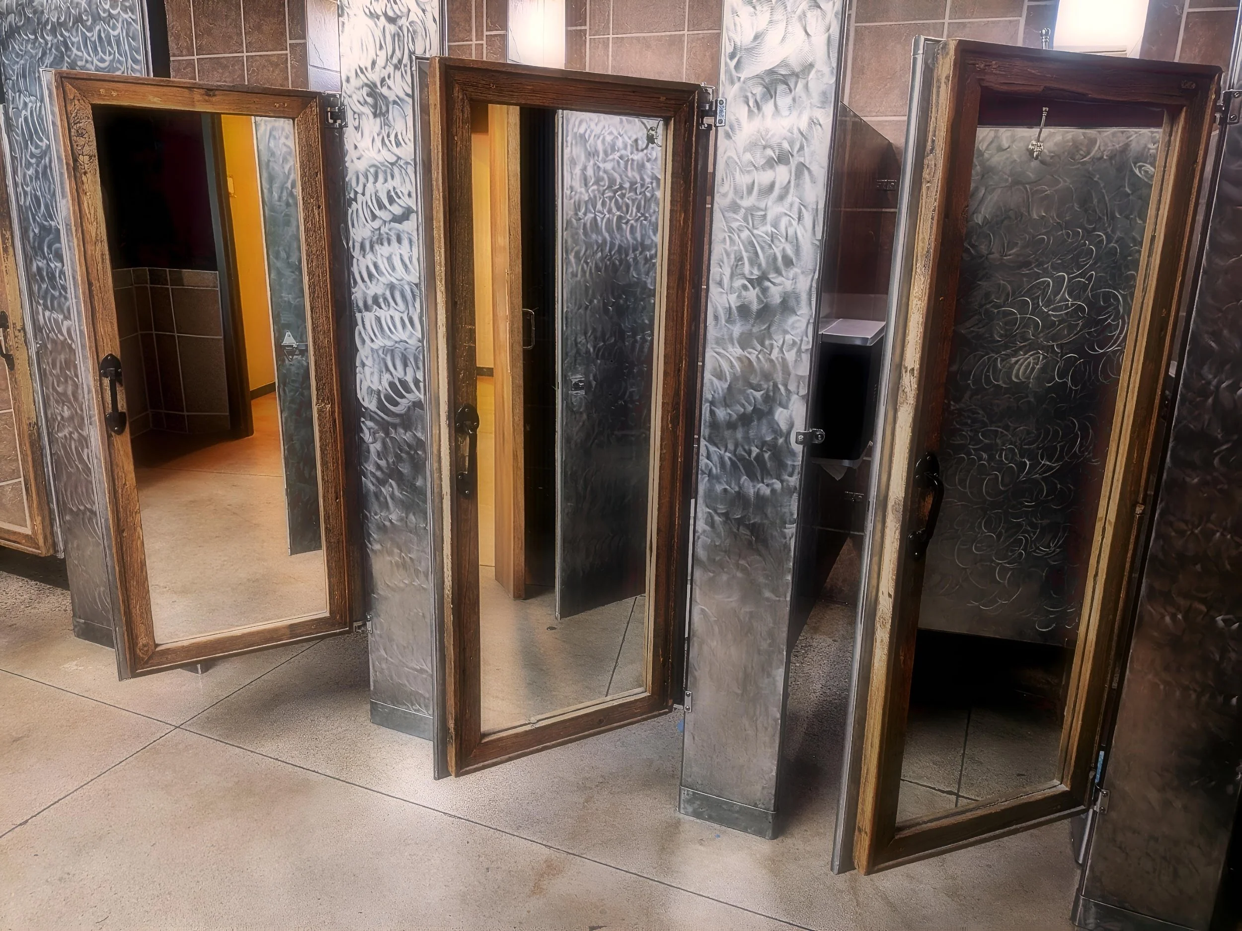 Unisex bathroom stalls with open wooden doors, metal frames, and a large mirror on the exterior of the leftmost stall. Tiled walls and concrete flooring are visible.