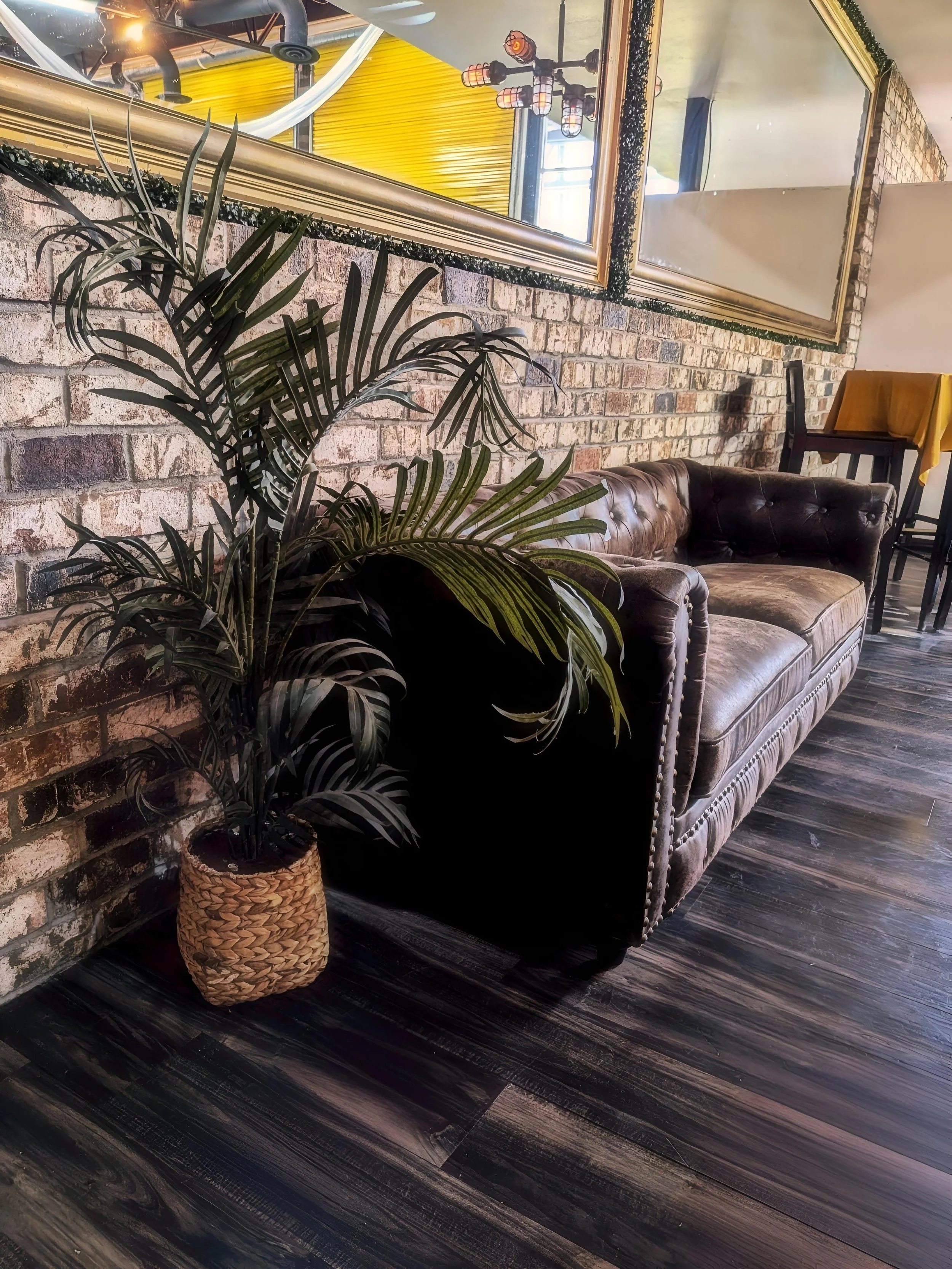 A living room corner with a vintage leather sofa, large potted plant, brick wall, and mirrored wall art.