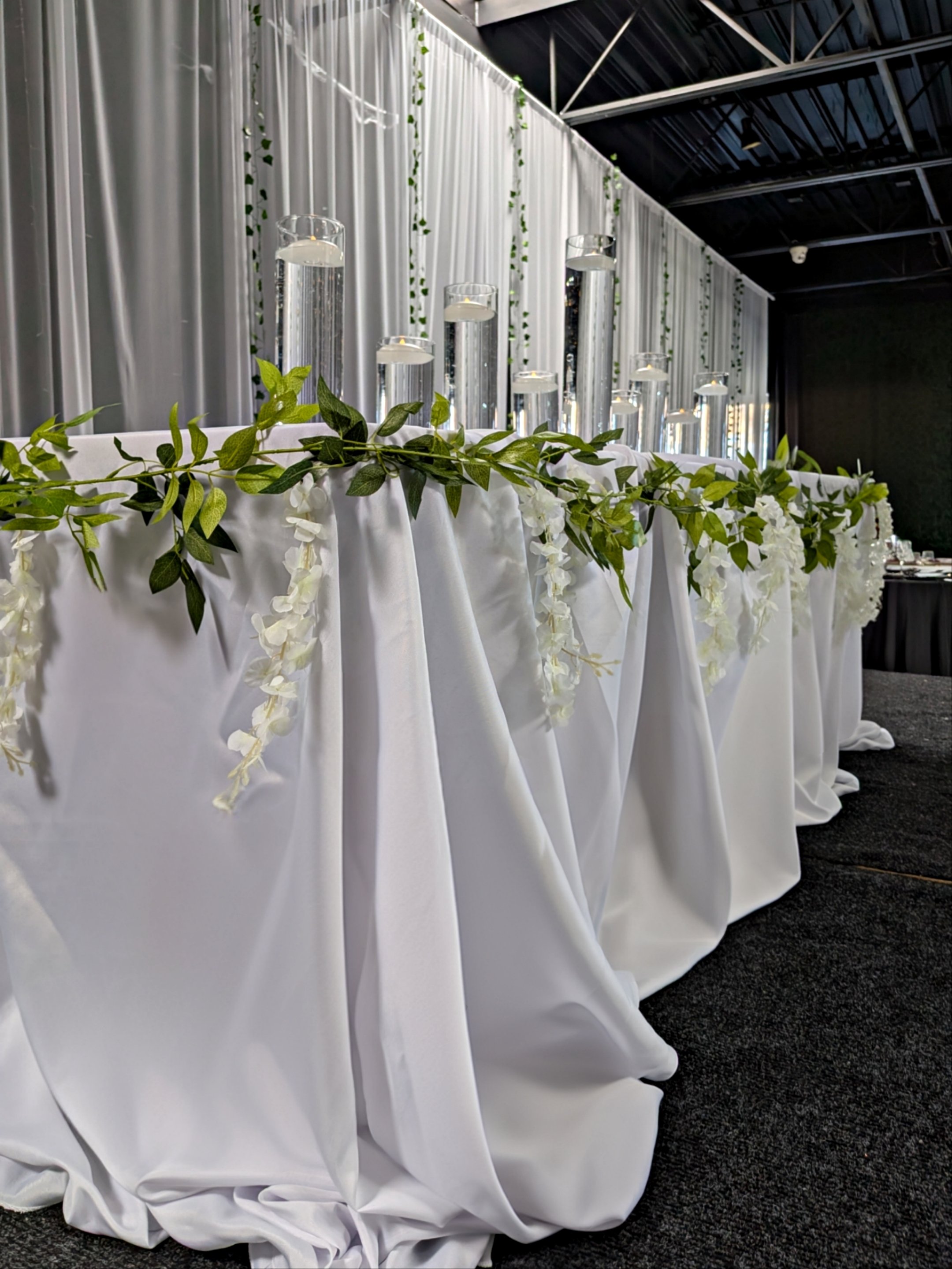 Elegant wedding or event table decorated with white draped fabric, greenery, and white flowers, with tall glass vases filled with water and floating candles, set against a backdrop of white curtains with hanging green vines.
