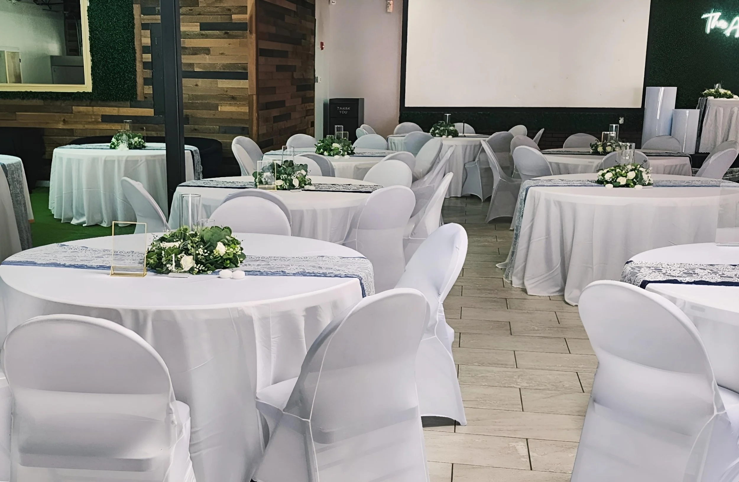 Indoor event space with round tables covered in white tablecloths, decorated with green and white floral centerpieces. White chairs surround the tables, and a large blank screen is on the wall in the background.