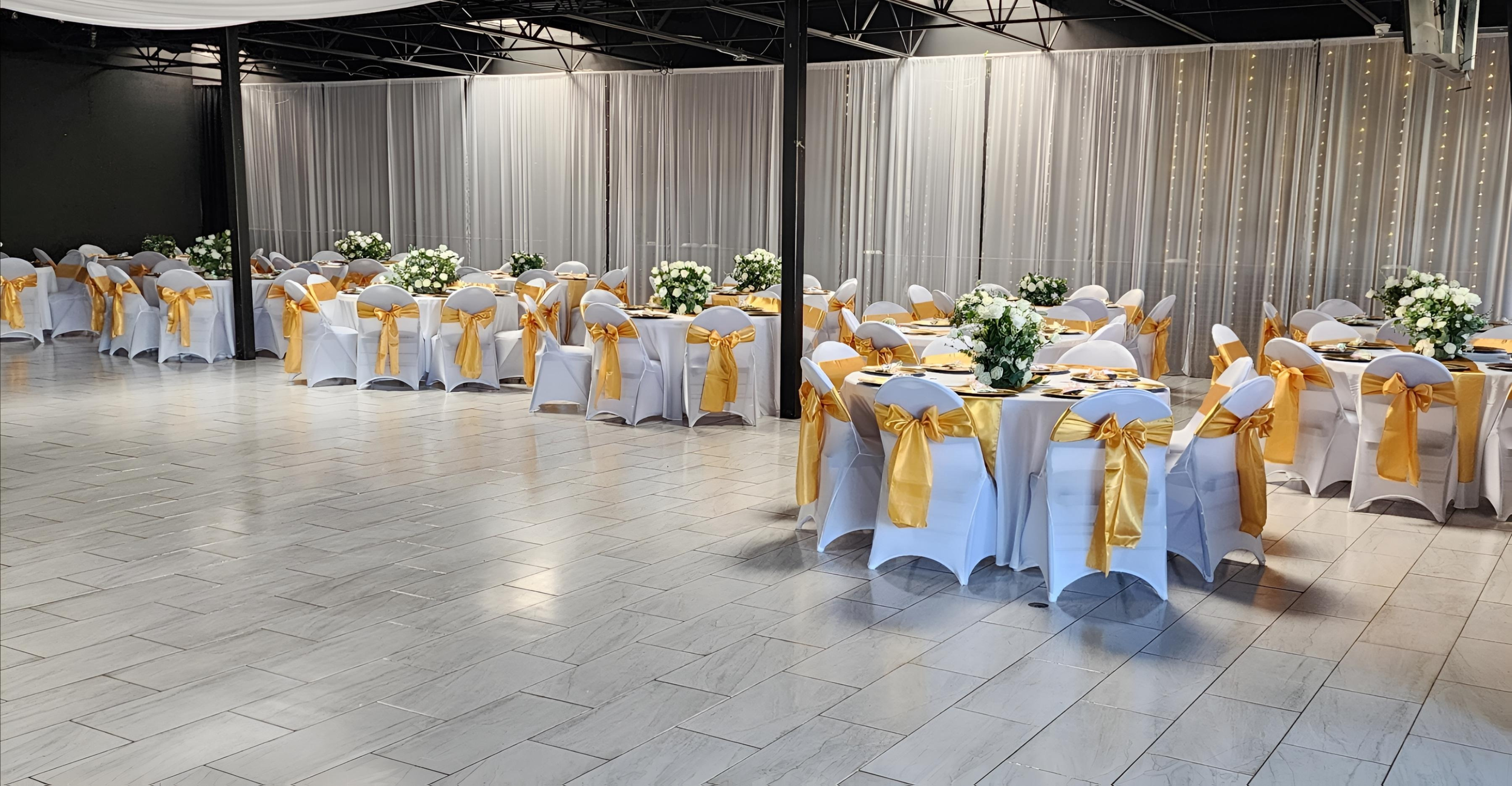 Elegant banquet hall with round tables covered in white tablecloths, decorated with large floral centerpieces. Each chair is draped with a white cover and tied with a gold sash. The hall has a black ceiling structure, light-colored tiled flooring, and sheer curtains with string lights forming the backdrop.