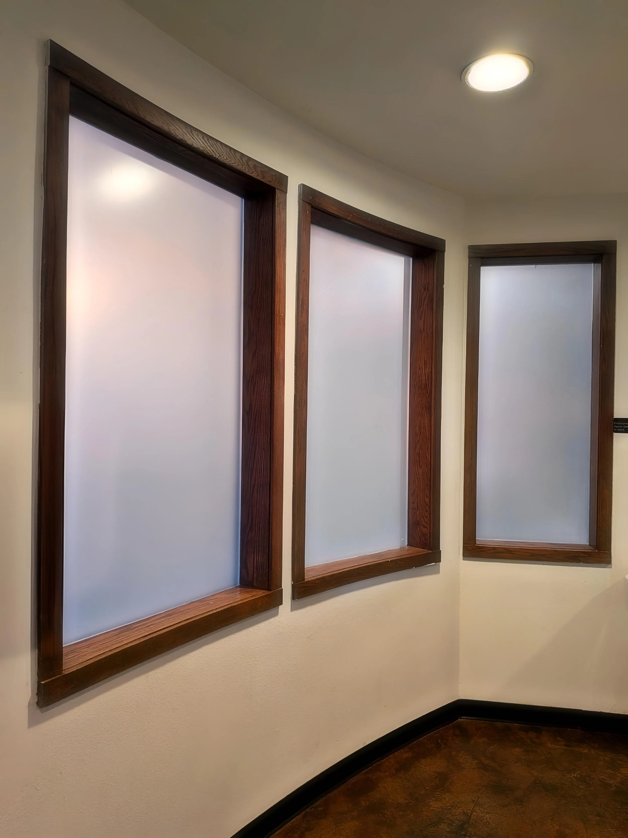 Three wall-mounted picture frames with frosted glass and dark wooden borders on a cream-colored wall in a room with a curved brown floor and ceiling lights.