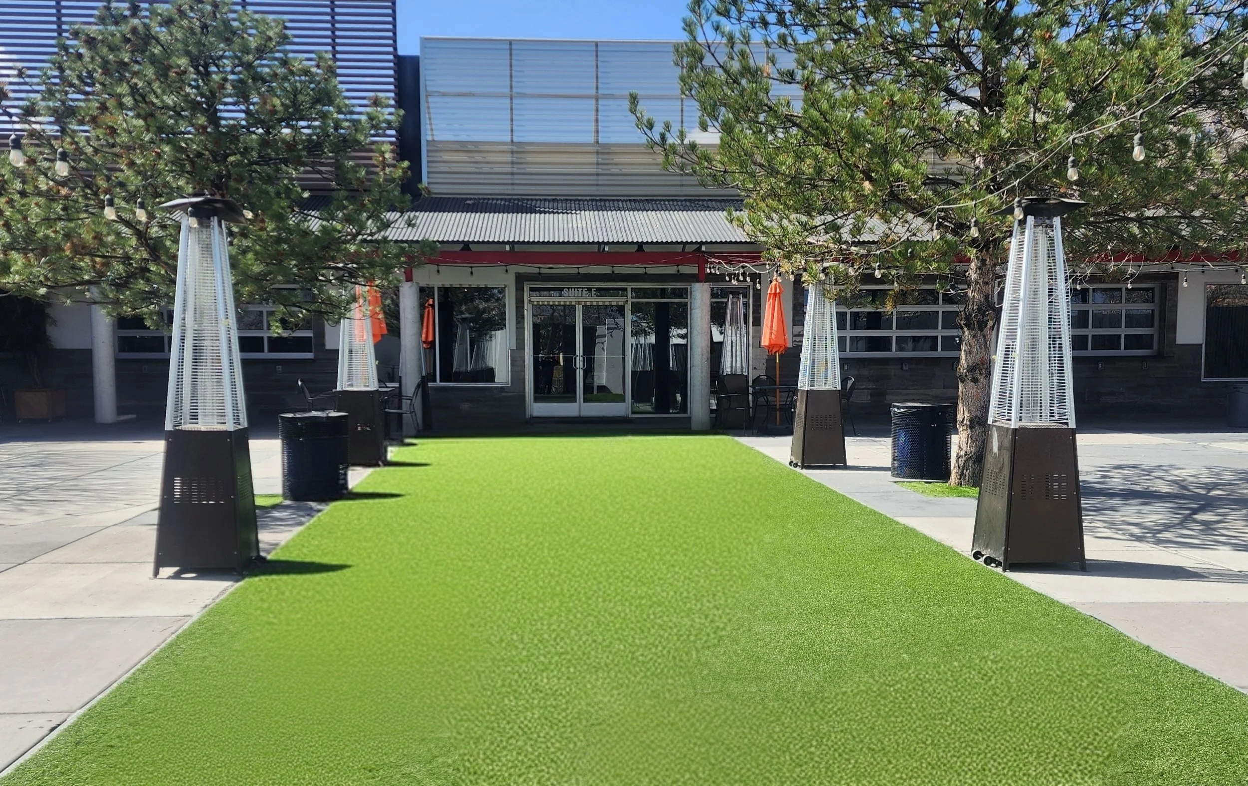 Outdoor patio area with artificial grass, string lights, trees, patio heaters, trash cans, and a building entrance with glass doors.