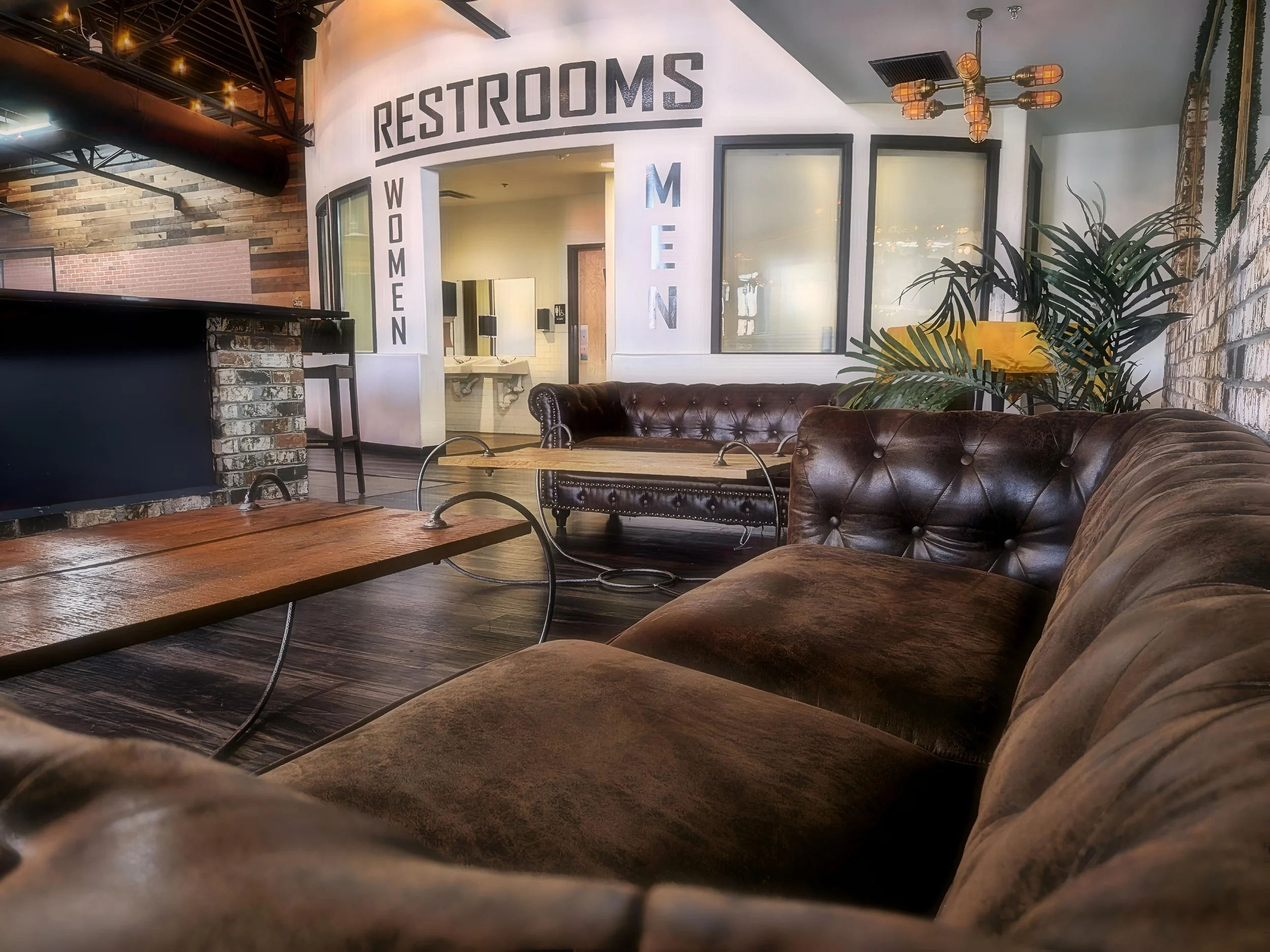 Interior of a cafe or lounge with leather sofas, wooden tables, potted plants, industrial-style lighting, exposed brick walls, and a sign indicating women's restrooms.
