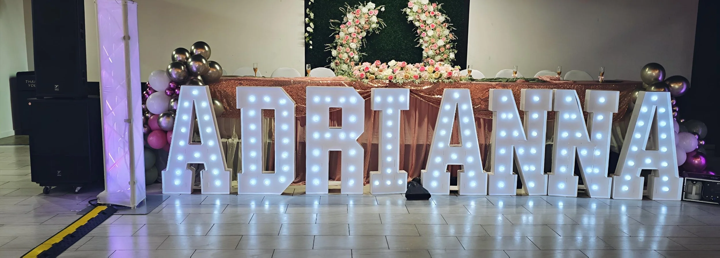 Large illuminated sign spelling 'ARLANA' with balloons and a floral backdrop at a decorated event table.