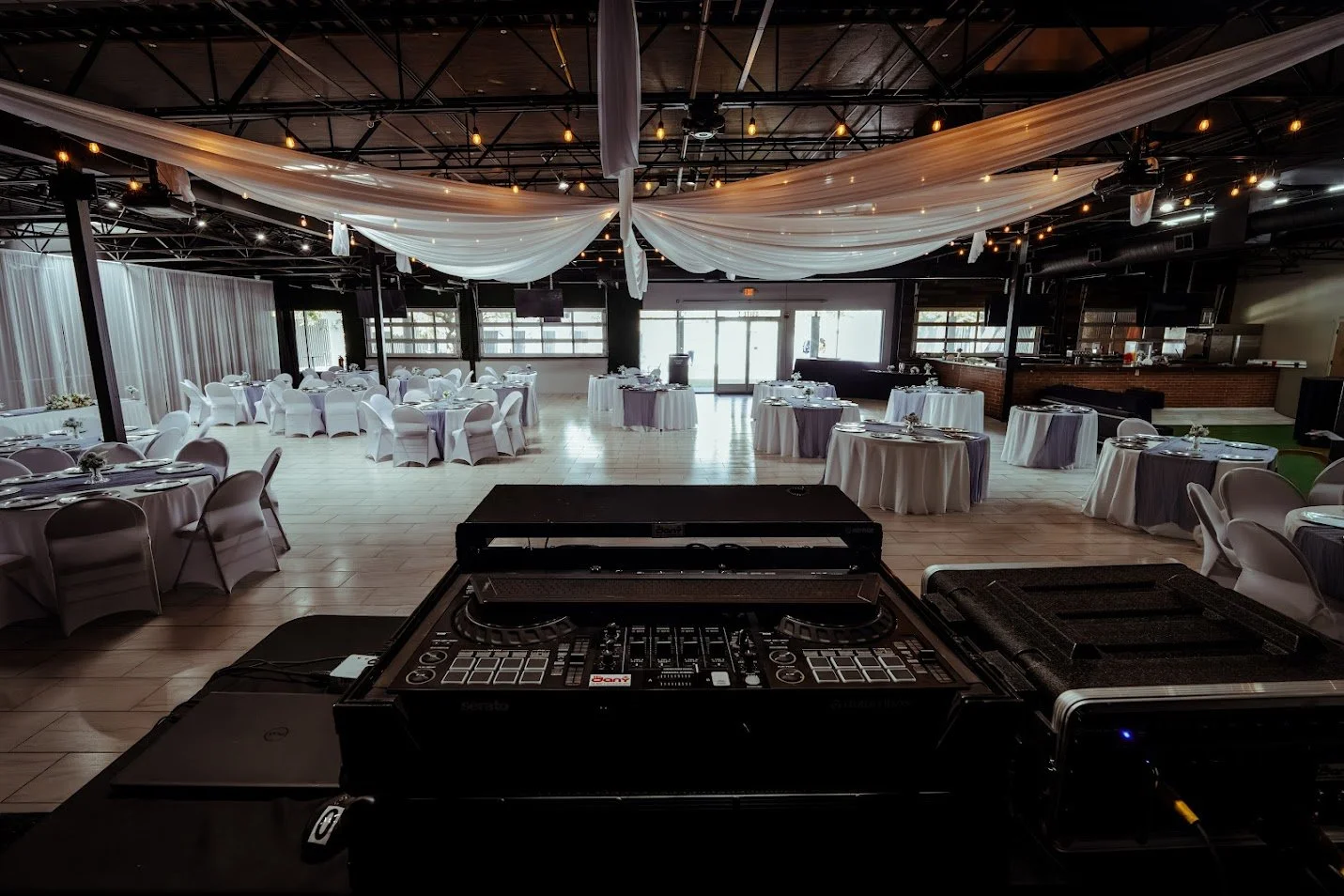 Event venue with round tables draped in white and purple tablecloths, set for a formal occasion, with draped fabric and string lights hanging from the ceiling, DJ equipment in the foreground, large windows, and a bar area in the background.