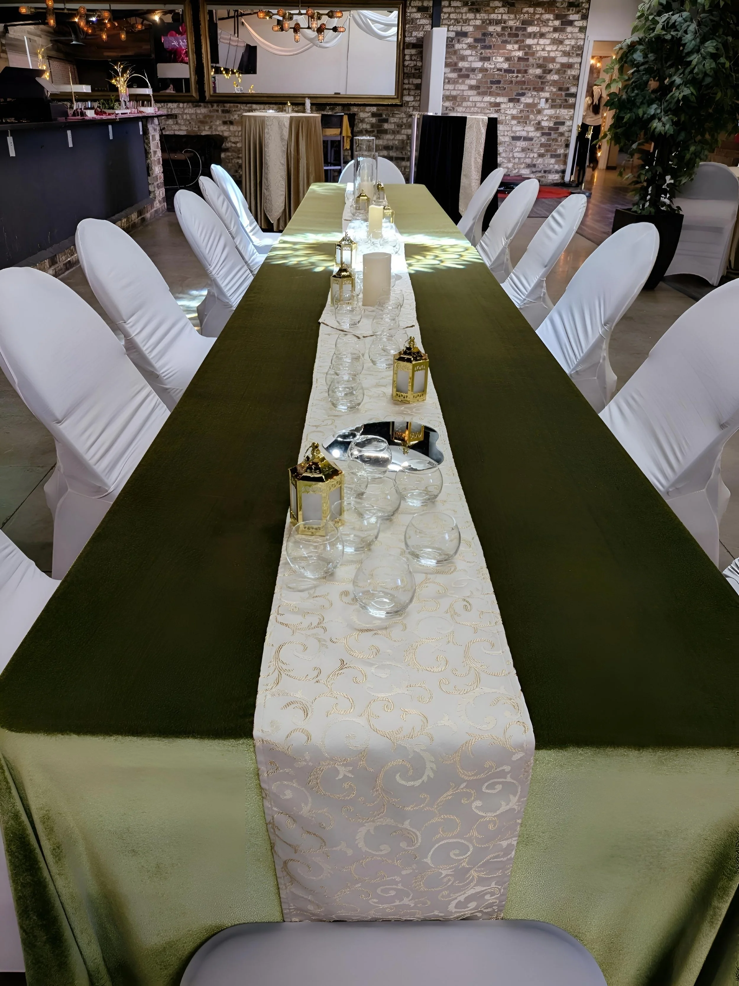 A long banquet table decorated with a green tablecloth and a white patterned runner. The table is set with glassware, small lanterns, and candles, with white chairs arranged around it in a venue with exposed brick walls and ambient lighting.