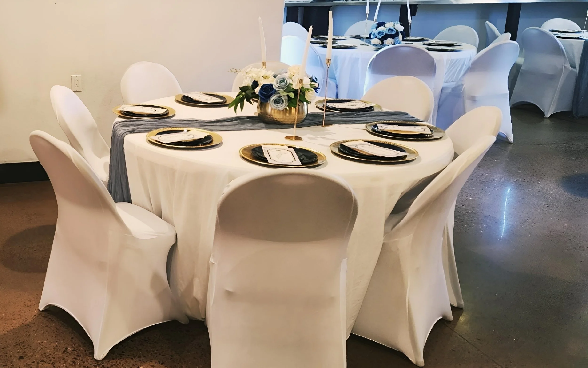 Round banquet table decorated with a dark gray runner, a floral centerpiece with white, blue, and gray flowers, and black, white, and gold place settings, in a formal event space.