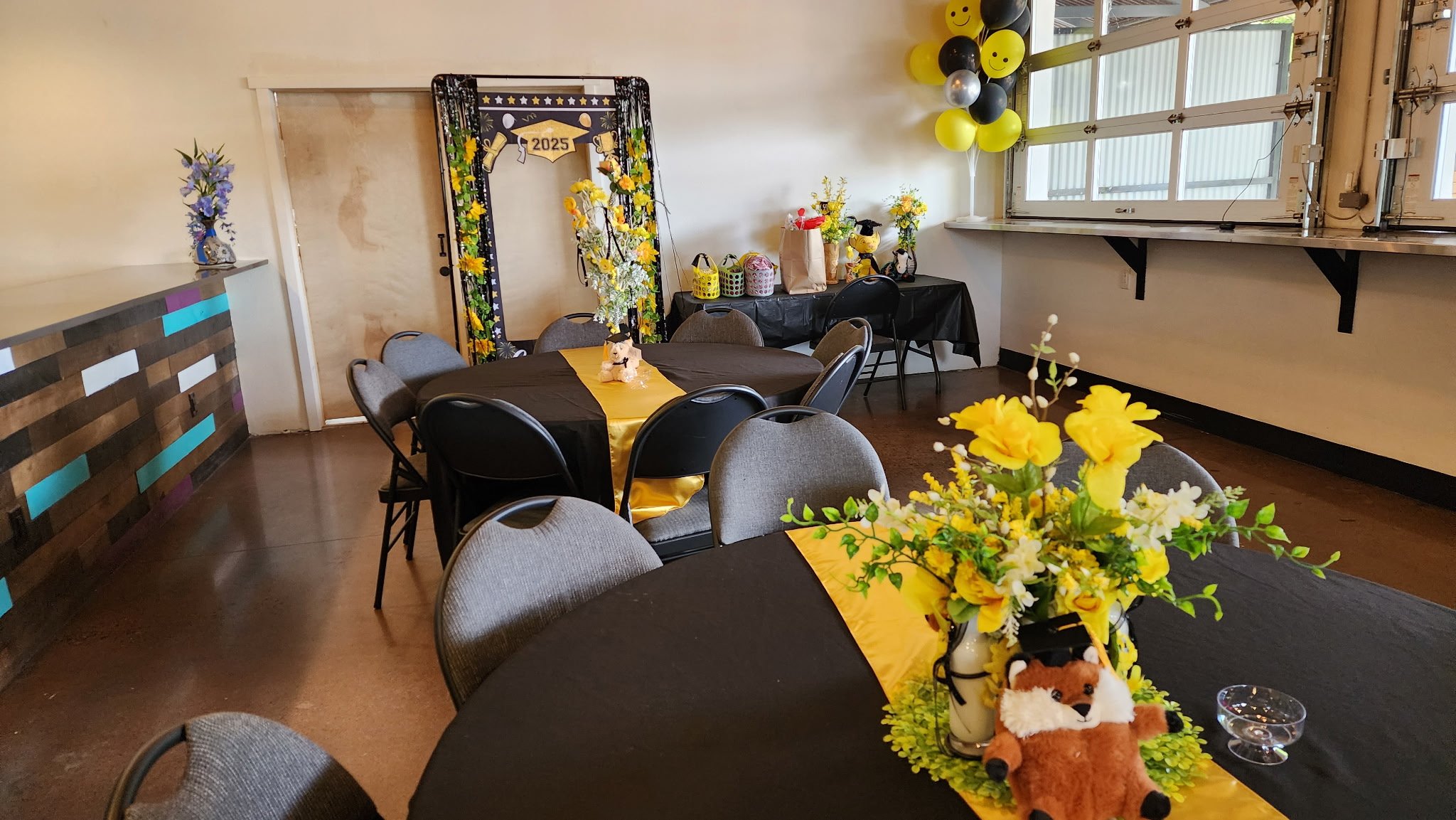 Party room decorated with yellow and black balloons, floral arrangements, and a graduation-themed backdrop with the year 2025. Tables are covered with black tablecloths, and one features a centerpiece with yellow flowers, a small fox plush, and yello