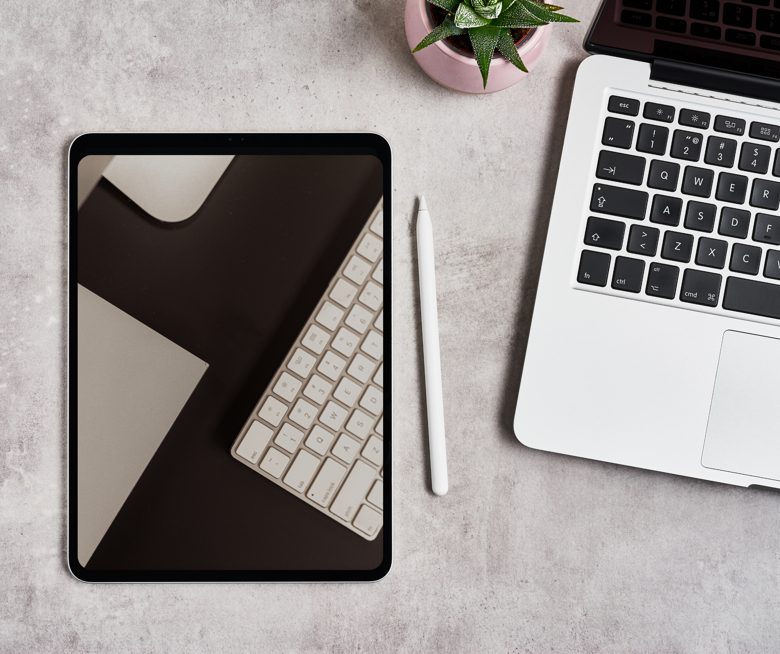 Mesa de trabajo con una tableta con pantalla apagada, teclado, bolígrafo blanco, una maceta pequeña con planta y una computadora portátil en un escritorio de color gris.