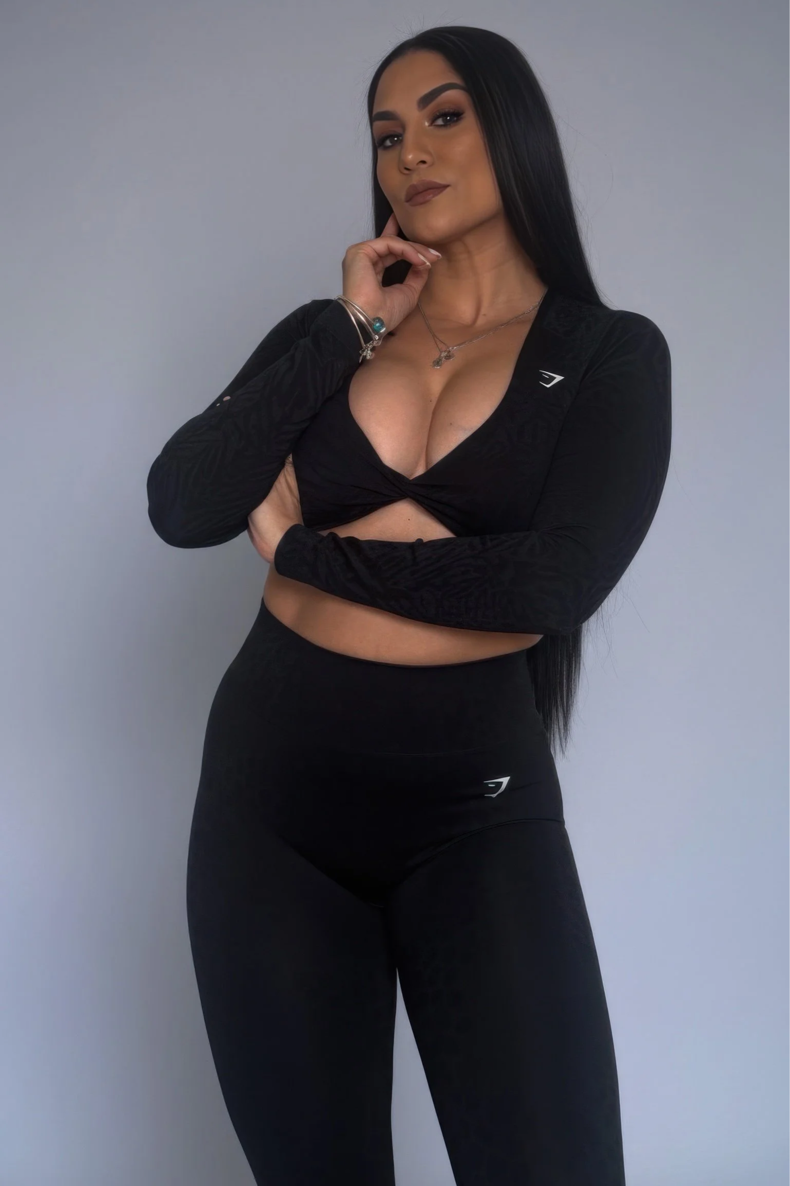 Mujer joven con cabello largo y oscuro, vestida con ropa deportiva negra, de pie contra pared gris, con una expresión confiada y pensativa.