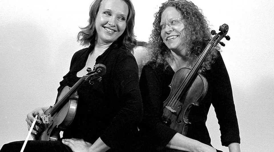 Two women smiling and holding violins, sitting back to back.