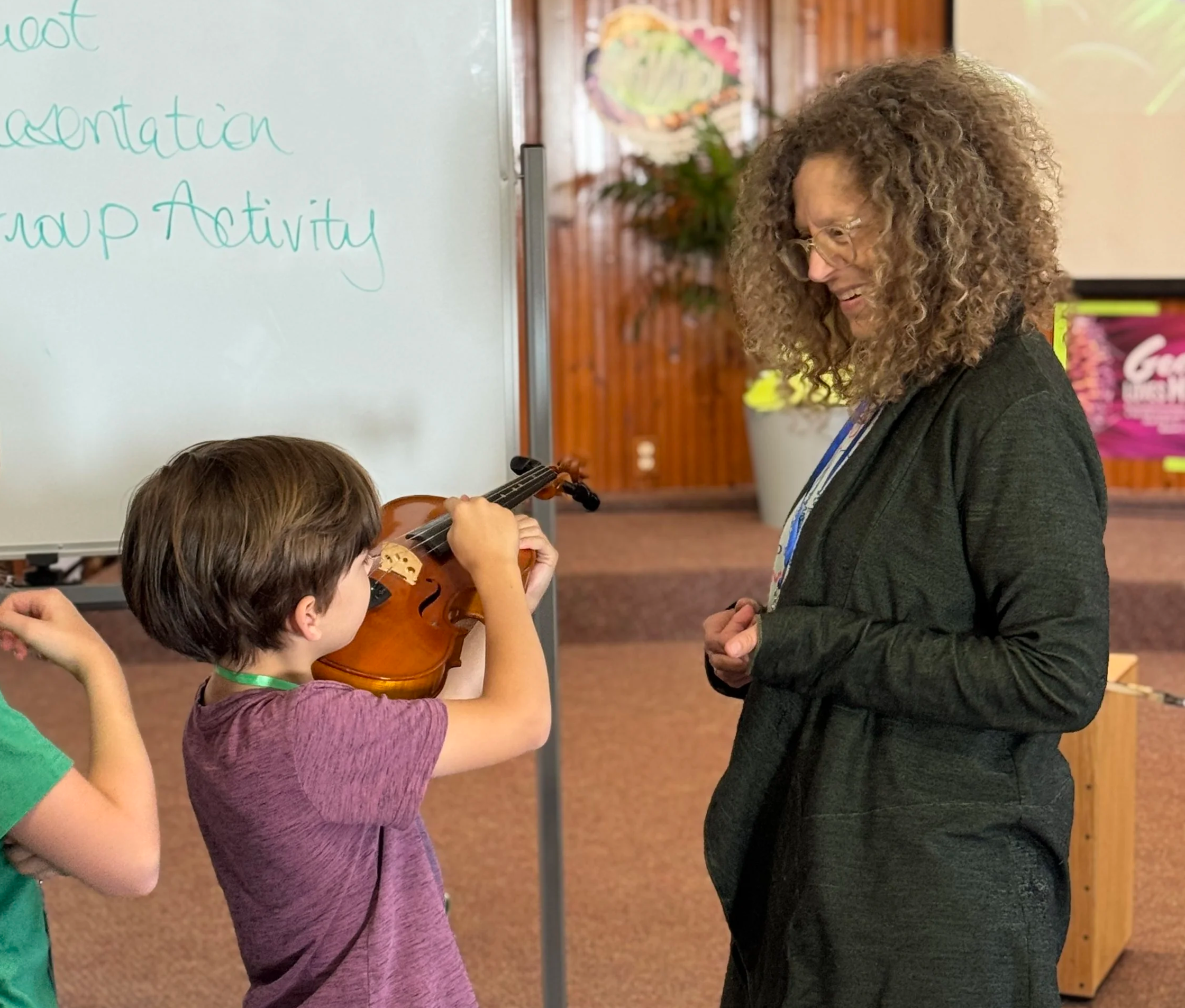 A young boy is playing a violin while a woman watches and smiles. Other children are partially visible. There is a whiteboard with writing and a potted plant in the background.