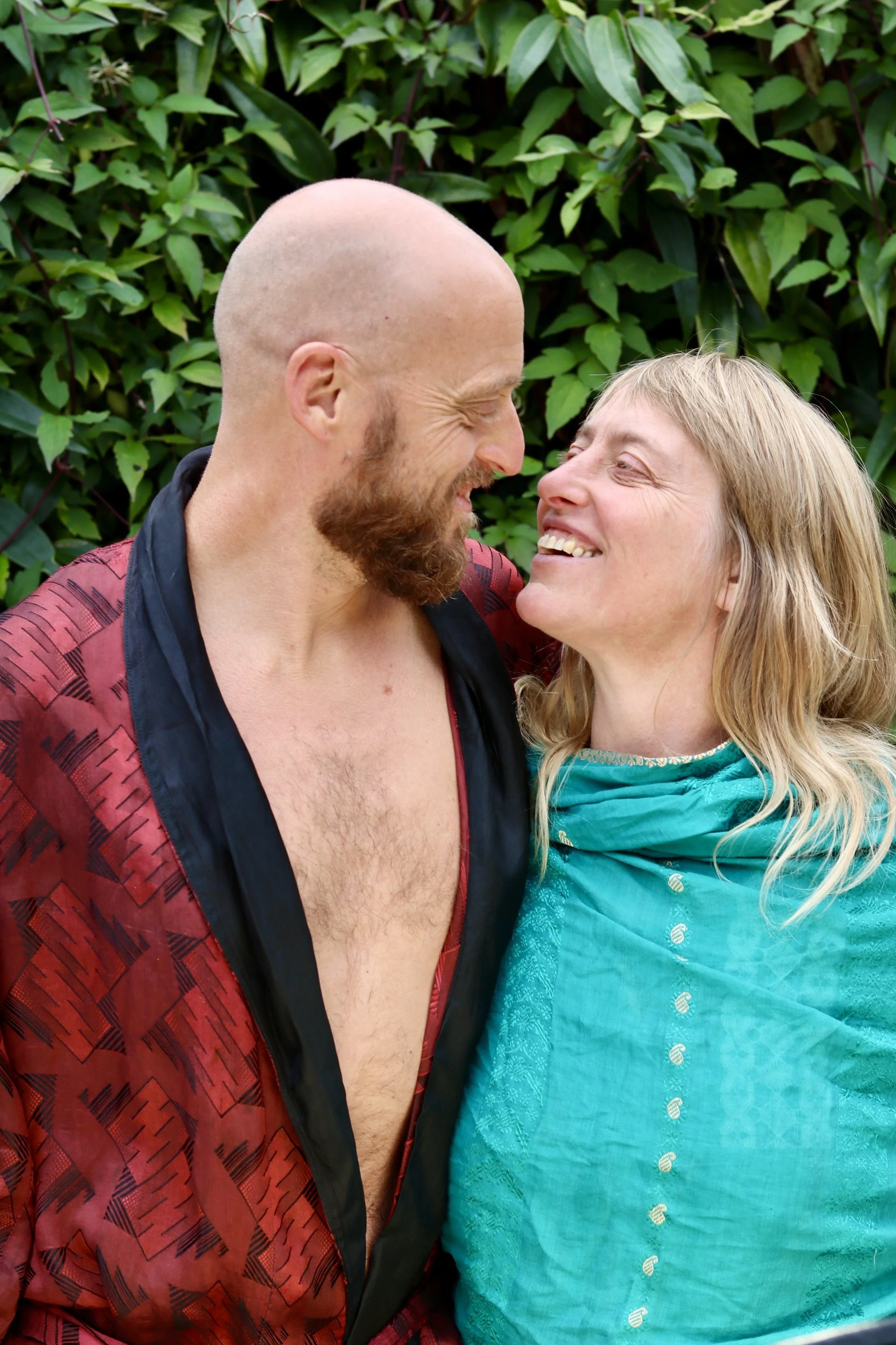 A man and woman smiling and looking at each other in front of green leafy background.