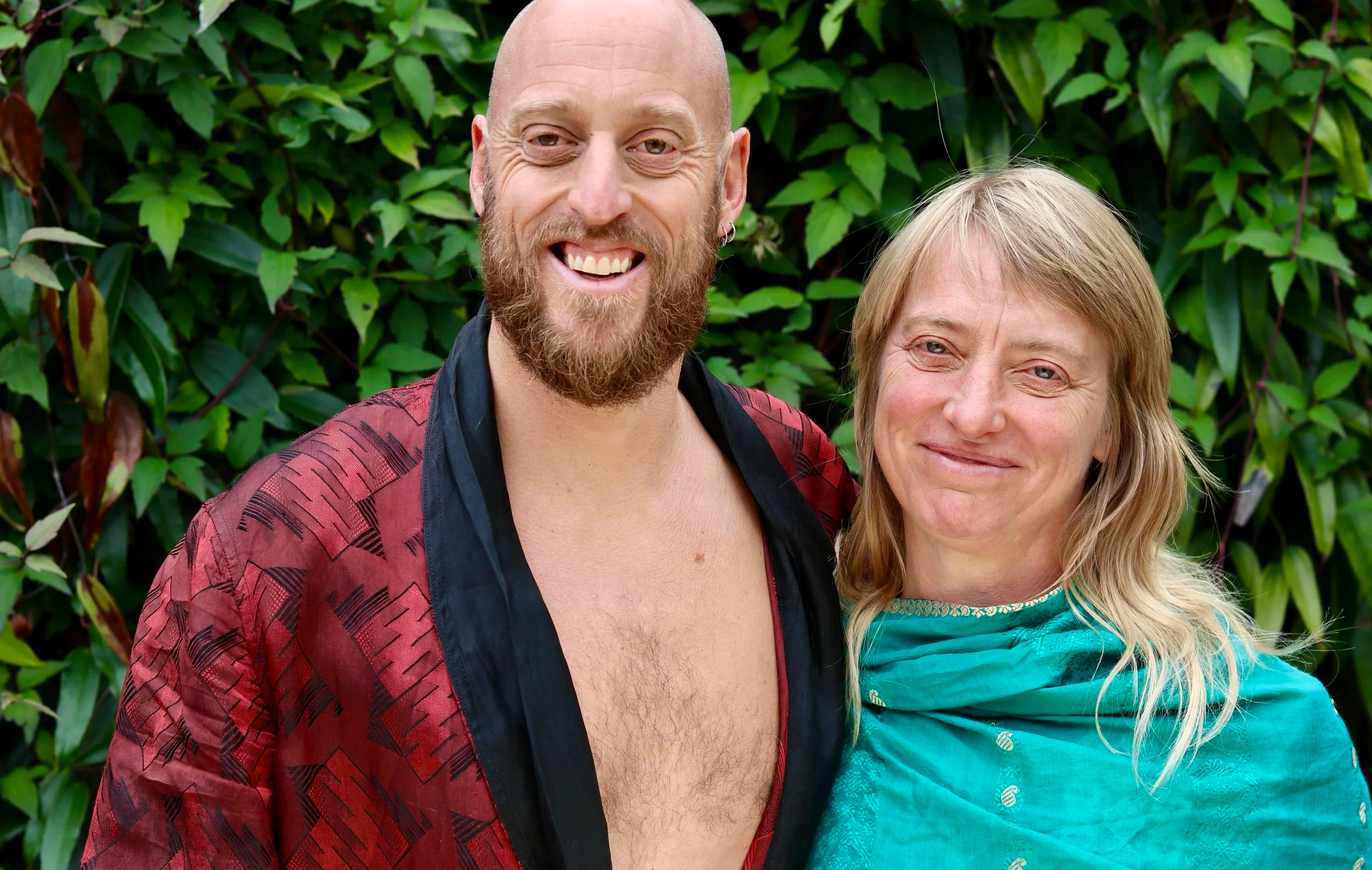 A smiling man with a beard and a woman with blonde hair standing outdoors in front of green foliage.