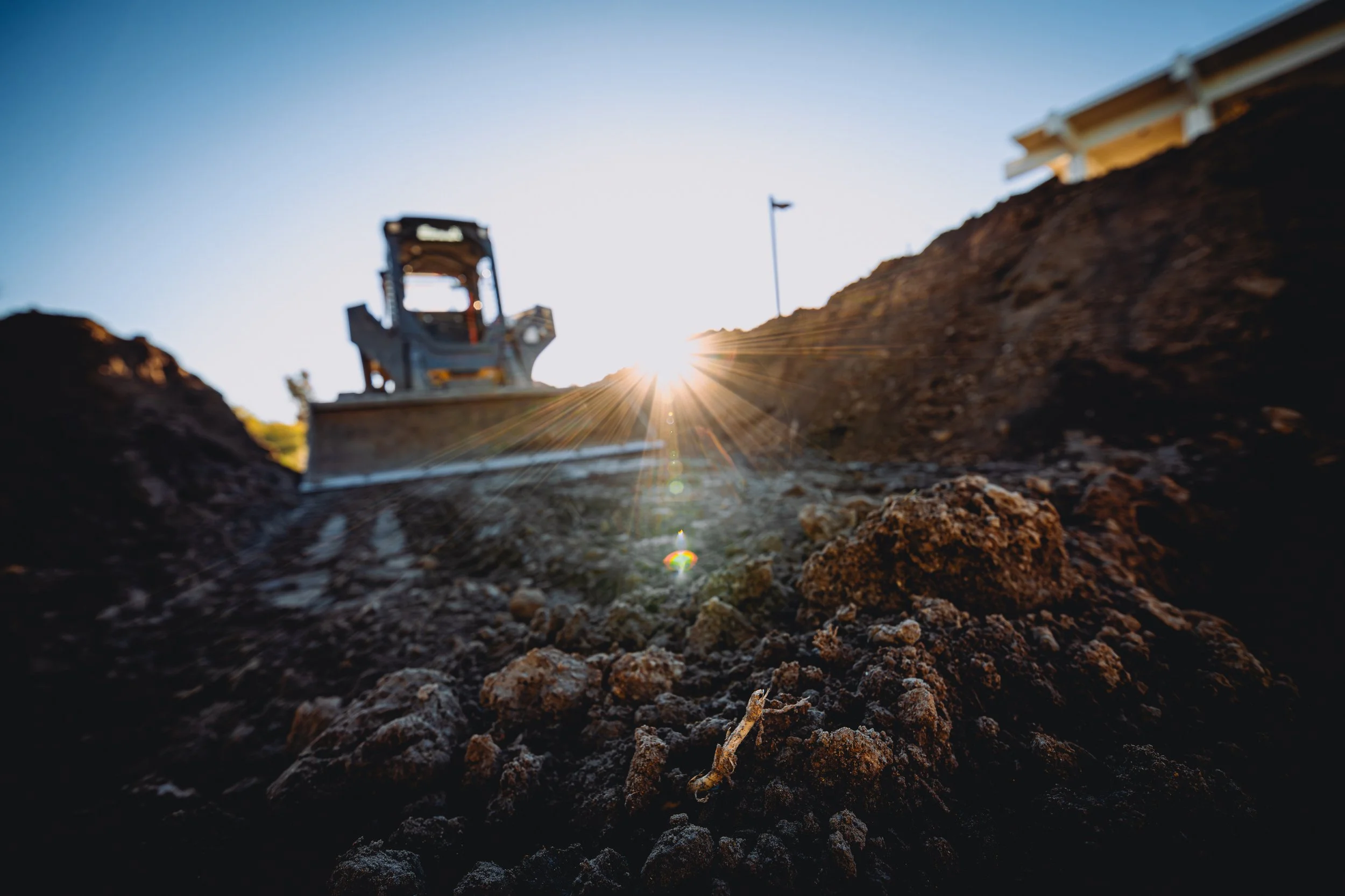 Construction equipment working on soil with the sun setting in the background, creating a lens flare.