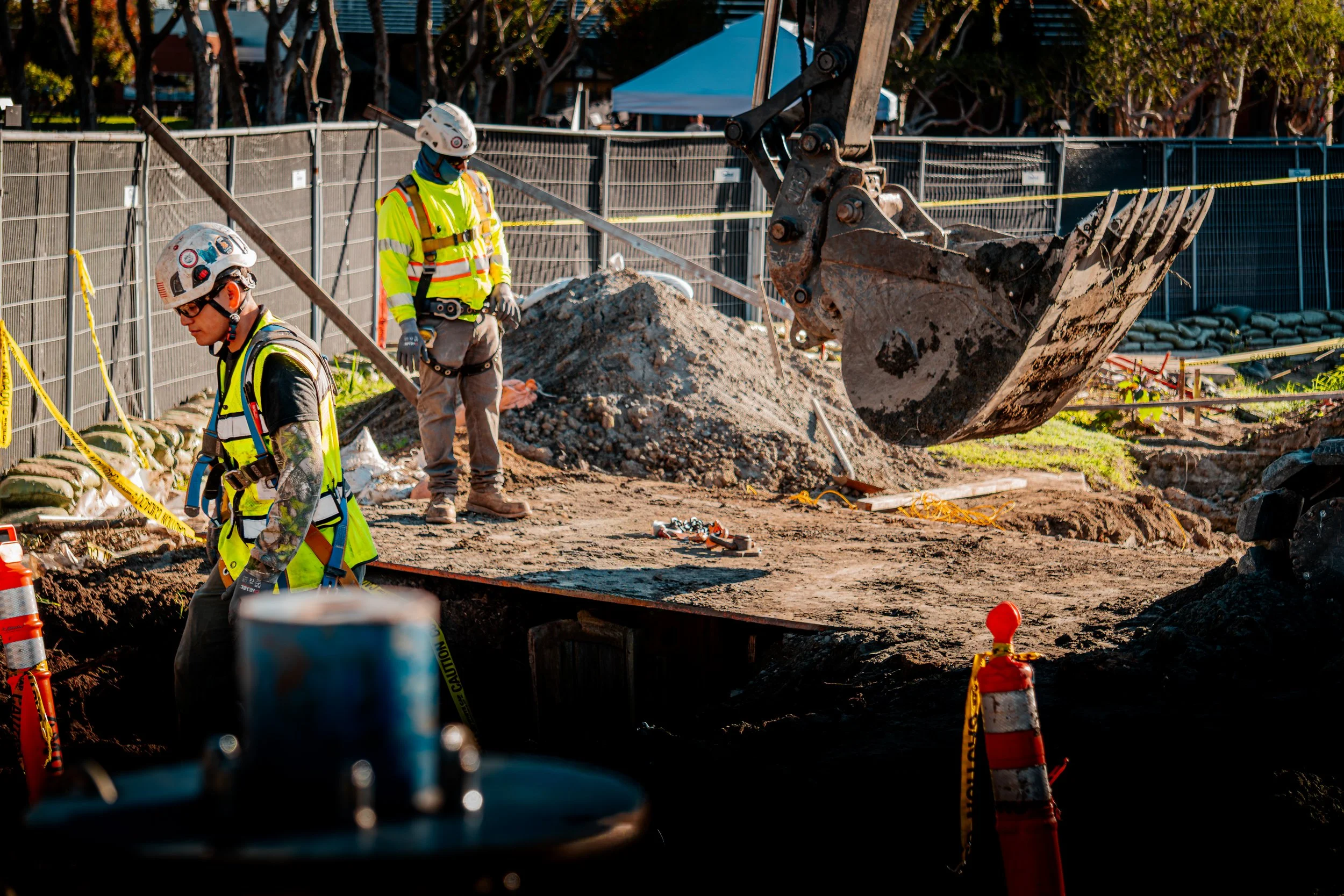 Construction workers in safety gear work at a construction site, with excavator and dirt mounds in background.