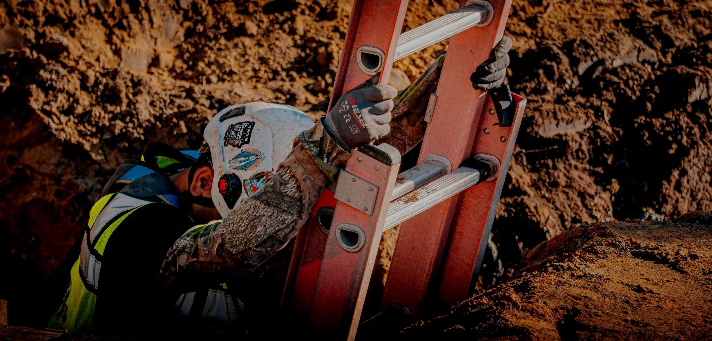 A construction worker wearing a white safety helmet with stickers, gloves, and a reflective vest is climbing a red ladder on a dirt construction site.