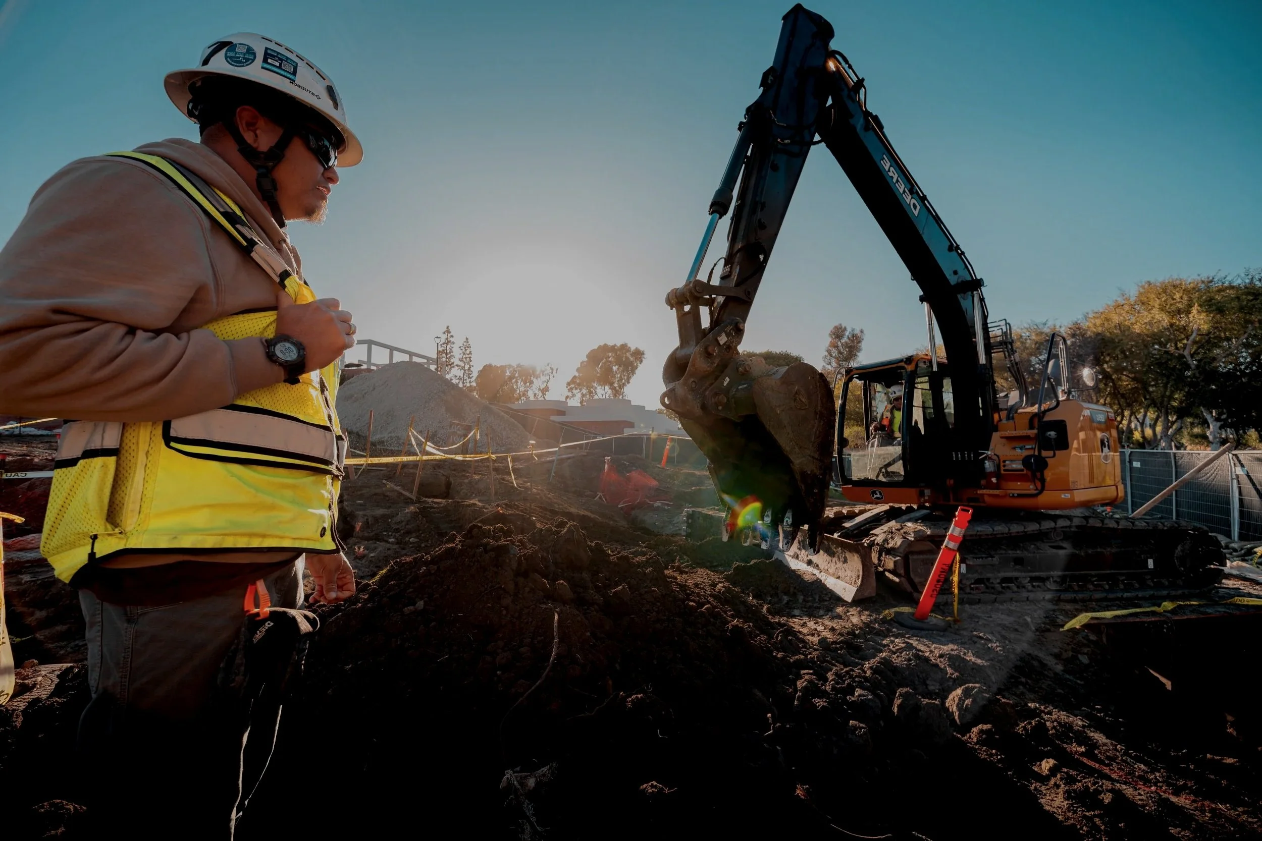Construction worker wearing a yellow safety vest, helmet, and sunglasses, standing next to a digging excavator on a construction site during sunset.