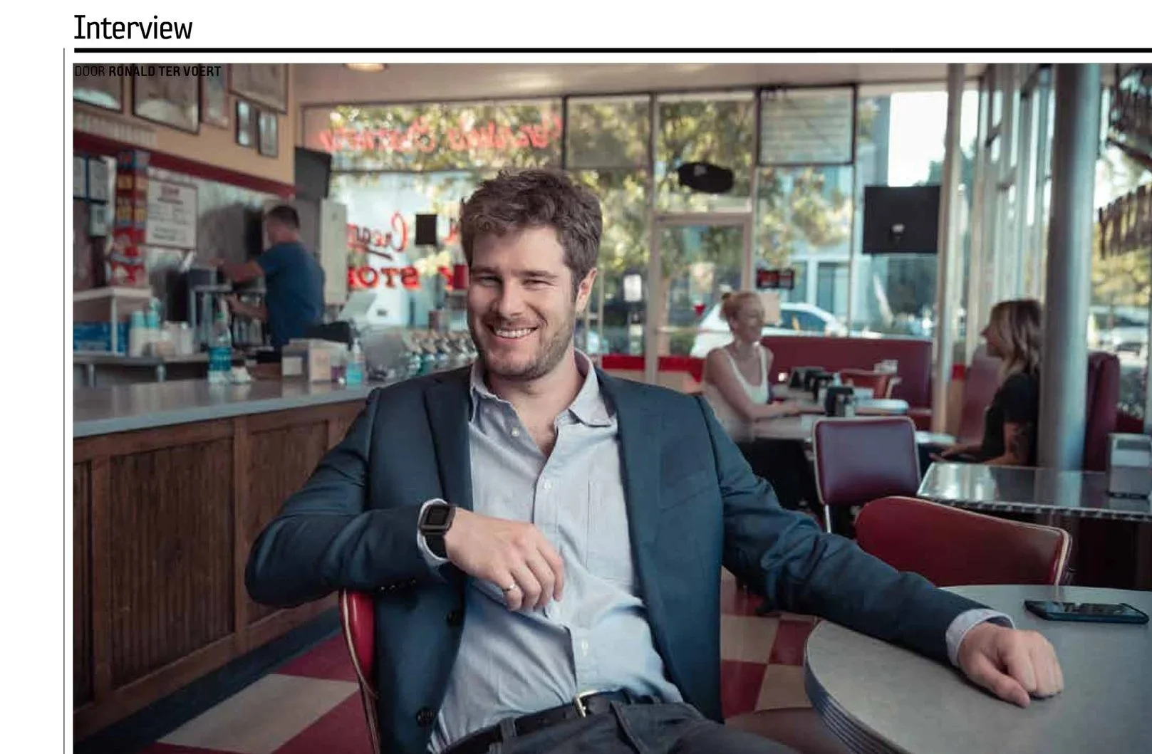 A man in a suit sitting at a table in a restaurant, smiling, with a watch on his wrist. There are other patrons and a worker in the background.