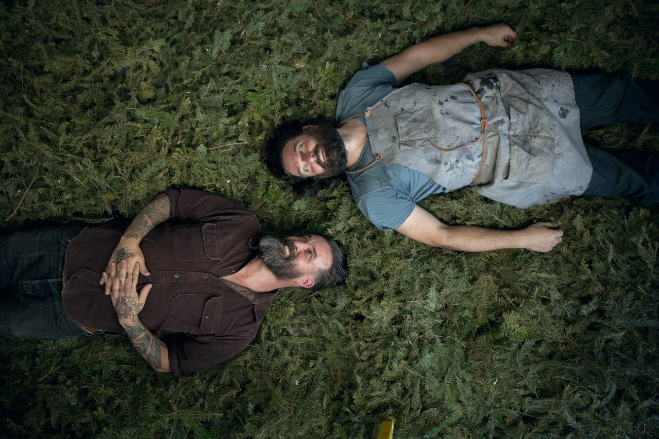Two men lie on a bed of marijuana plants, one wearing a dark shirt with tattoos, the other in a stained apron and t-shirt, both smiling and looking at each other.