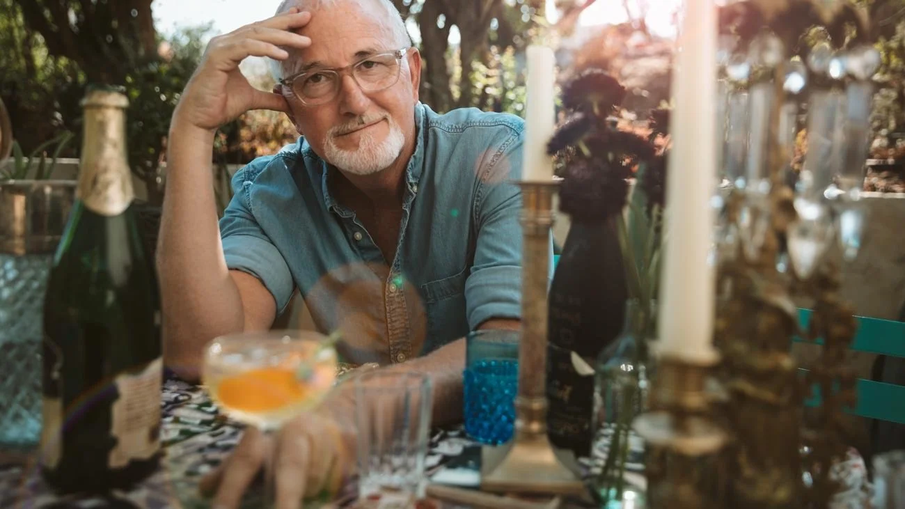 An older man with glasses and a short white beard seated outdoors at a table decorated with candles, bottles, and glasses, looking at the camera with a relaxed expression.