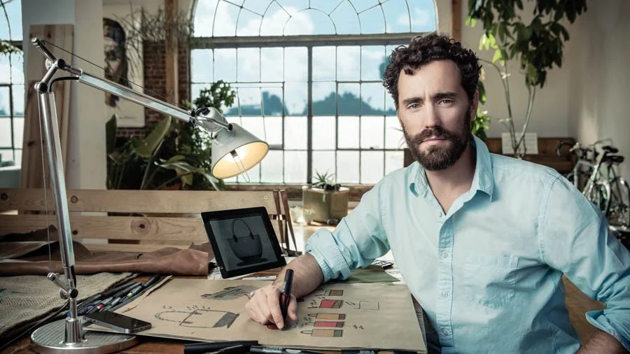 A man with dark curly hair and a beard sitting at a desk in a bright industrial-style room, working on a design with a pen. The desk has a lamp, a tablet, and several design sketches. Large windows and green plants are in the background.