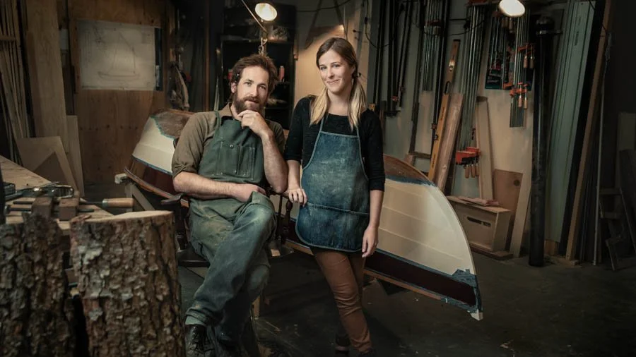 A man and a woman in a woodworking shop, standing in front of a boat under construction, surrounded by woodworking tools and materials.