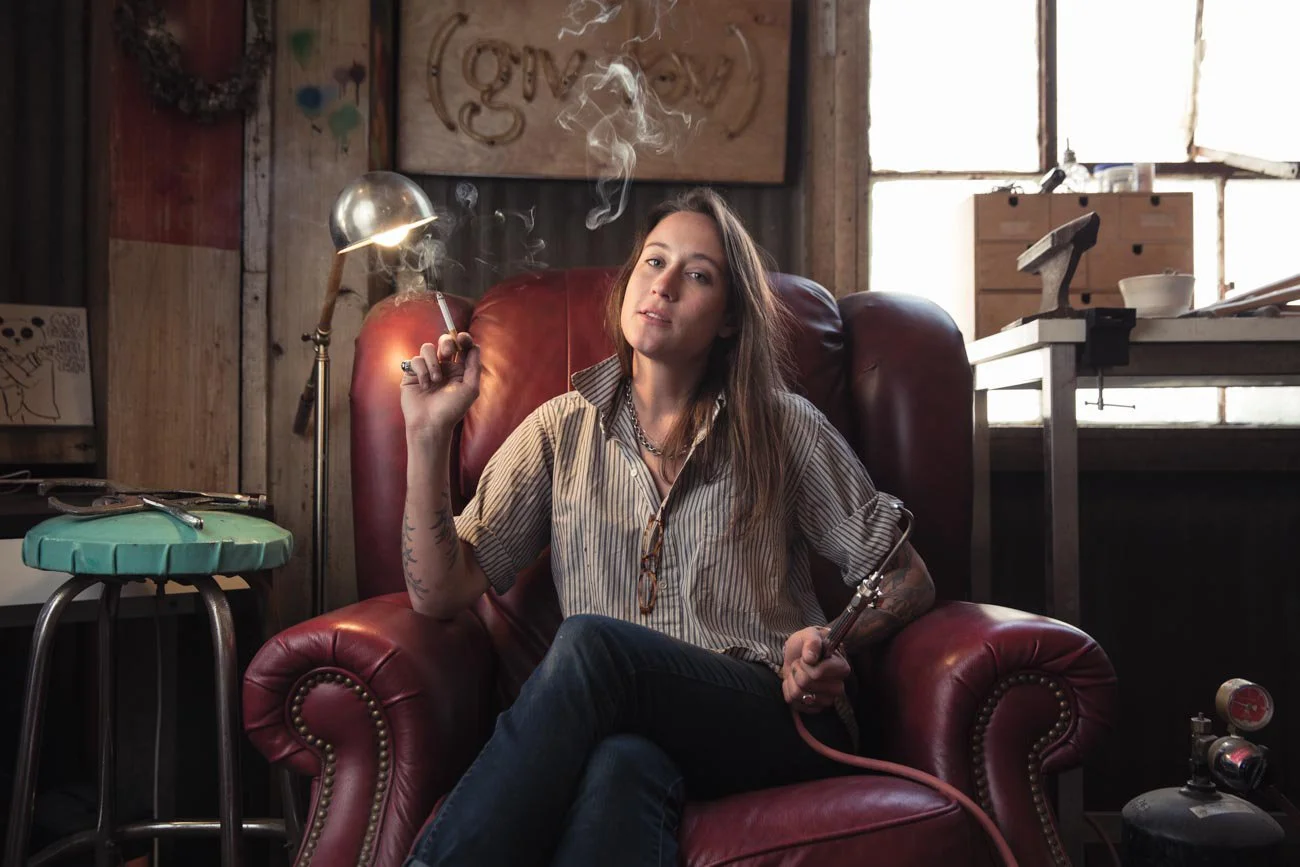 Young woman with tattoos sitting in a red leather armchair, smoking a cigarette, with a hookah hose in hand, in a rustic room with wooden walls and shelves, behind her a window with sunlight streaming in.