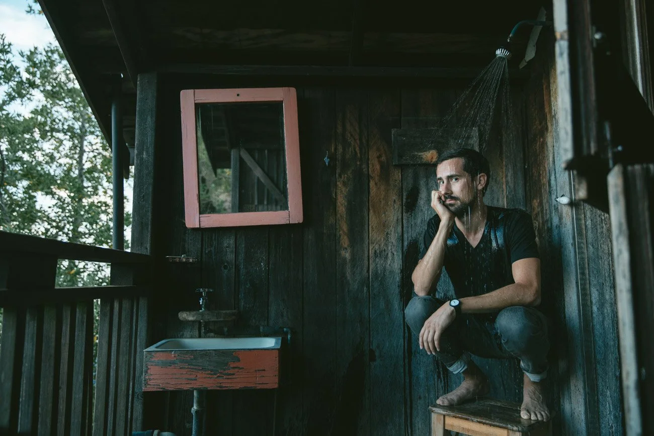 A man sitting in a shower area with wooden walls, rain water coming from the showerhead, near a small sink and a mirror, outdoors with trees visible outside.