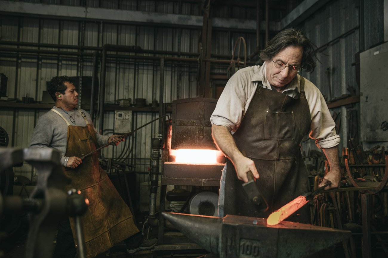 Two blacksmiths working in a forge, one pouring molten metal while the other hammers hot metal on an anvil.