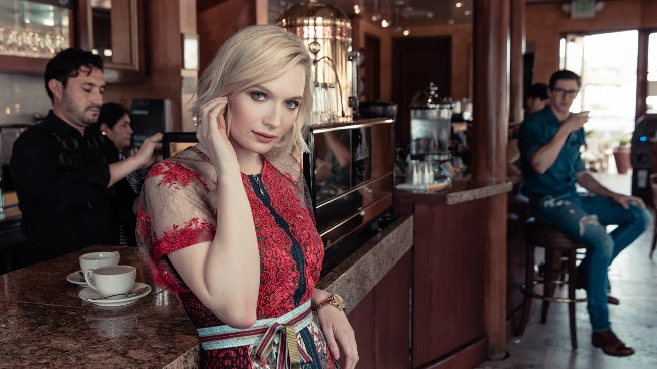 A blonde woman in a red and black lace dress posing at a cafe counter, with a coffee machine and cups behind her, and people in the background.